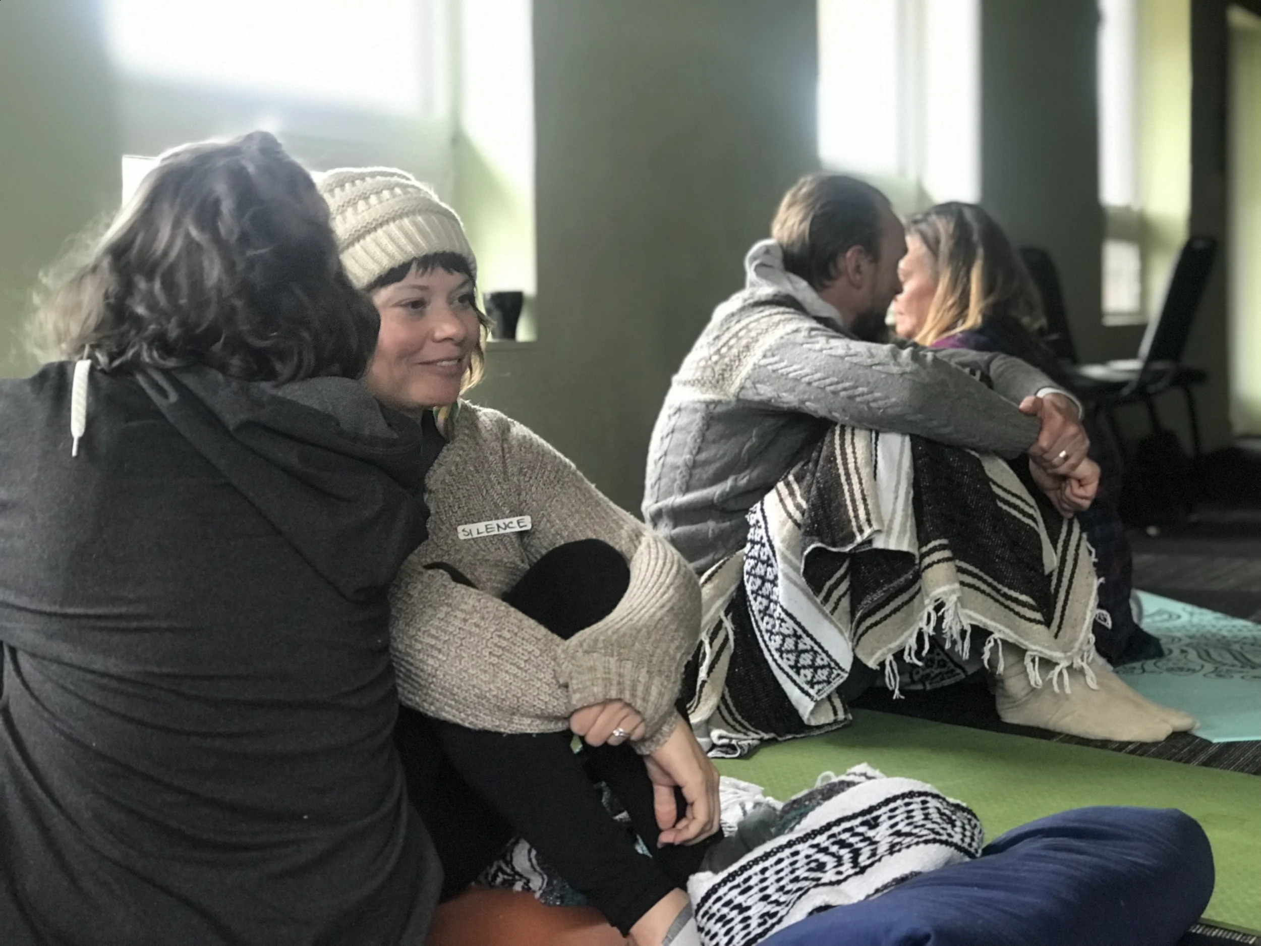 People sitting on the floor with their eyes closed, possibly participating in a meditation or mindfulness session. One person in the foreground has a label that reads 'Silence' on their sleeve.
