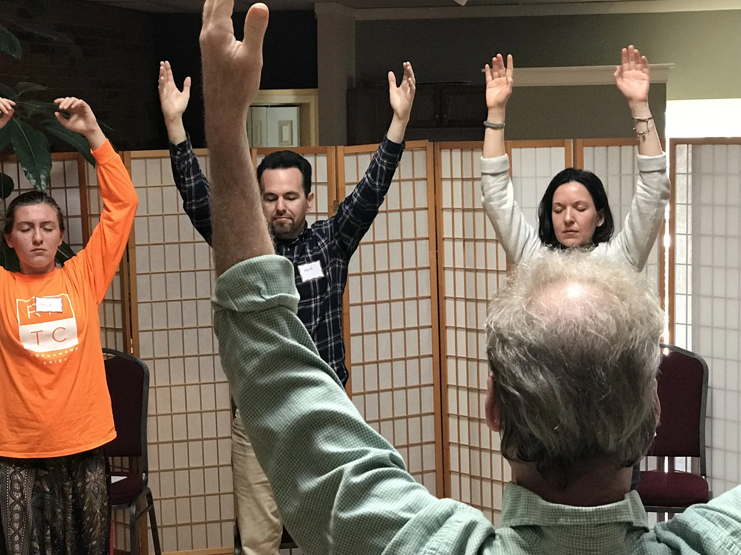 People participating in a meditation or mindfulness session with hands raised, sitting in front of a folding screen.