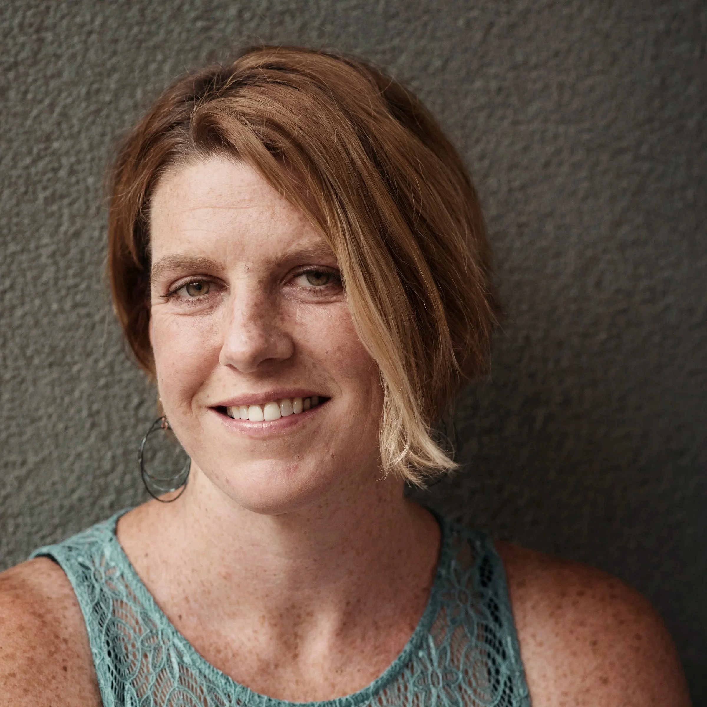 A woman with short, wavy, reddish-brown hair smiling while standing against a gray textured wall.