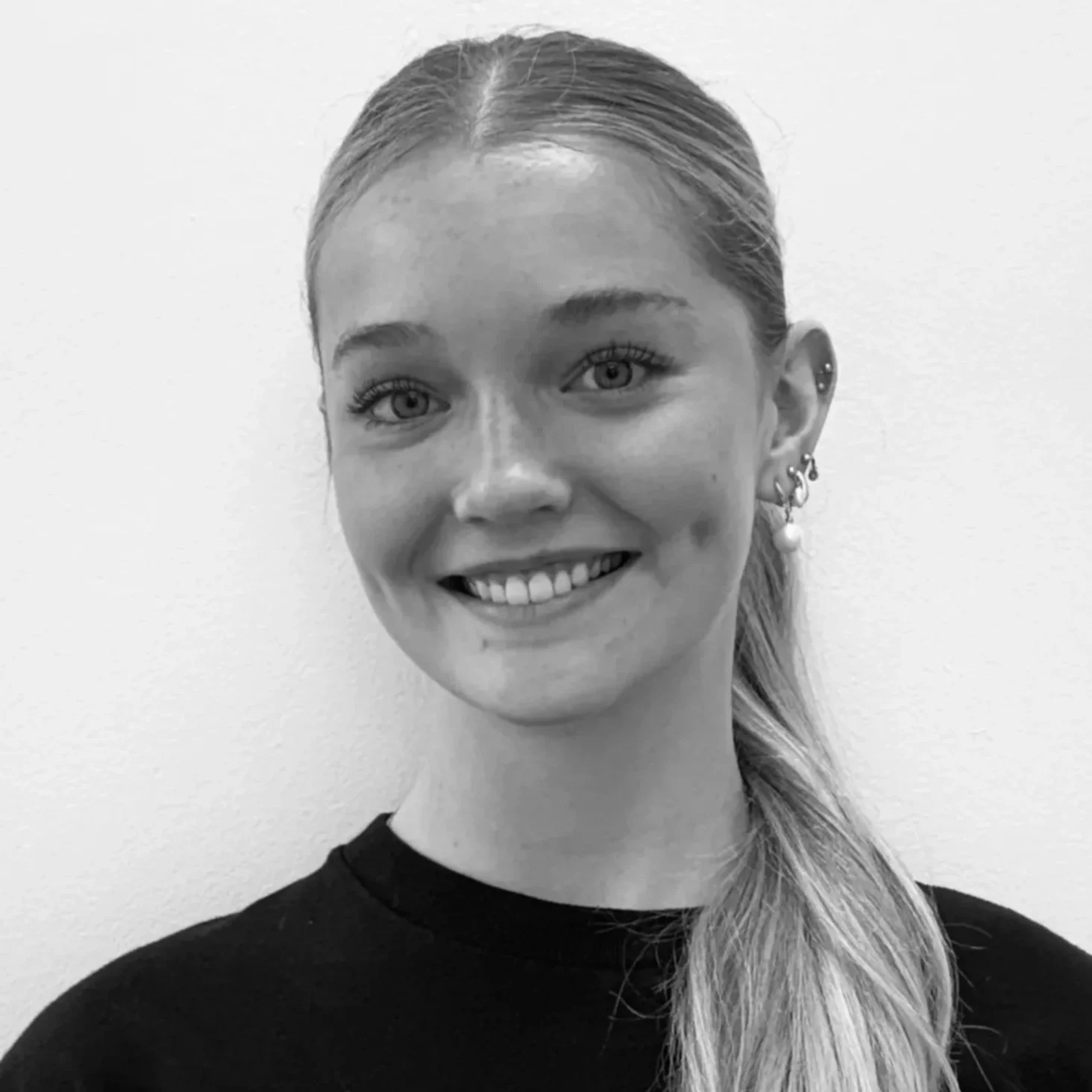 Black and white photo of a young woman with light-colored hair in a side braid, smiling, wearing earrings and a dark shirt, standing against a plain light background.