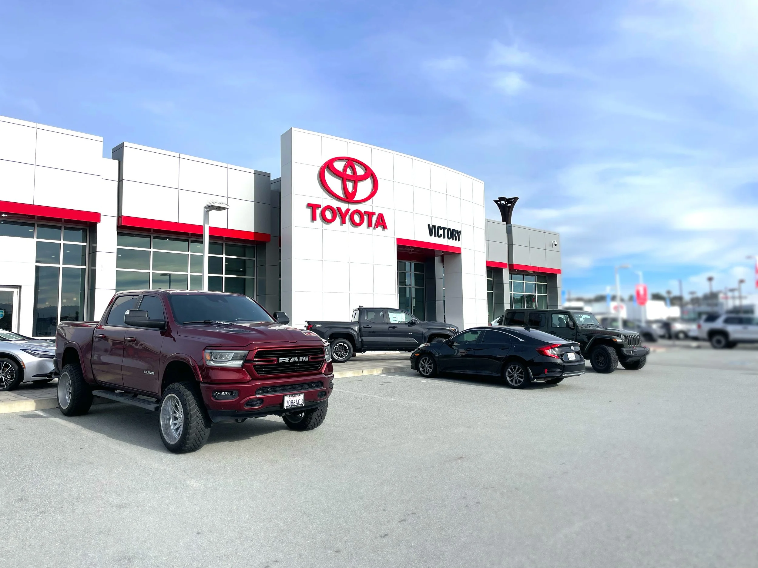 Toyota dealership with various new cars parked in front, including a maroon Ram truck, a black pickup, and a black sedan, under a partly cloudy sky.