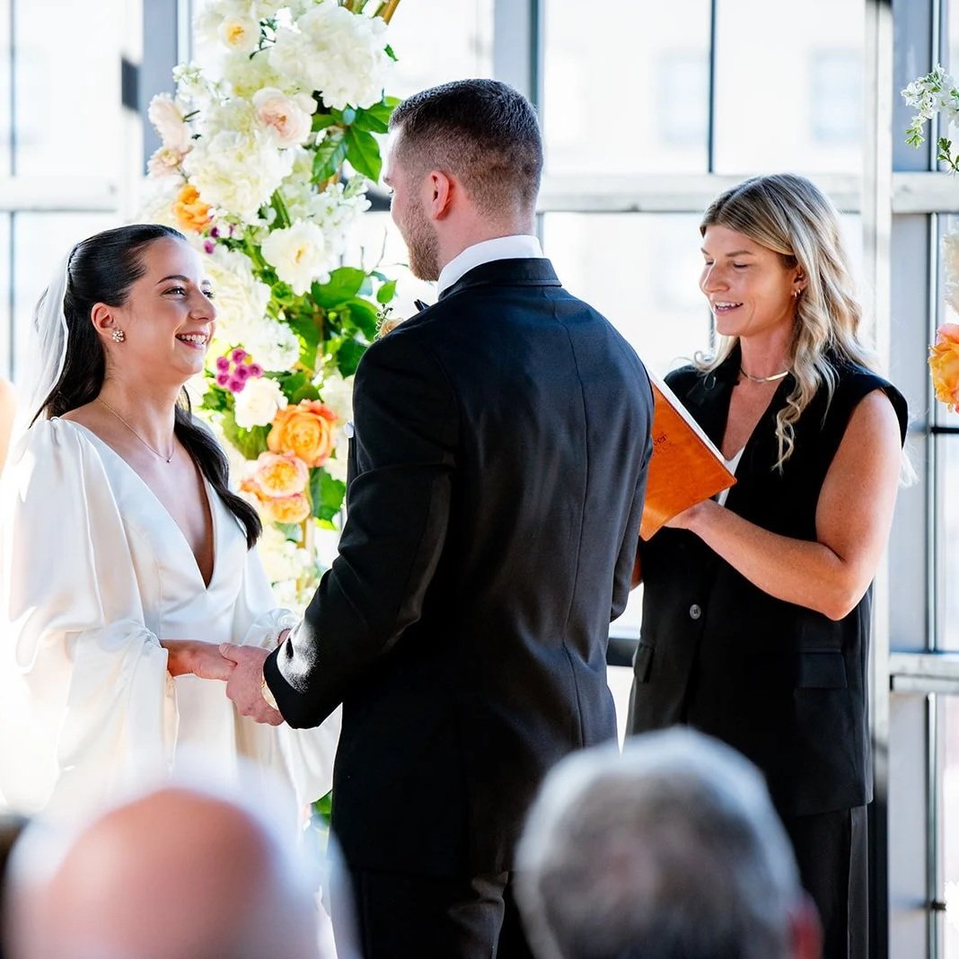 A couple getting married at an indoor wedding ceremony with a female officiant holding a orange folder, standing in front of a floral backdrop with white and pink flowers, during the daytime.