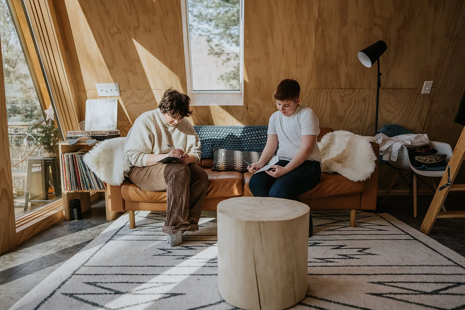 Two people sitting on a cozy leather couch in a room with wooden walls, writing or drawing on notebooks, with sunlight coming through windows. There are blankets, a small table, and a bookshelf nearby.