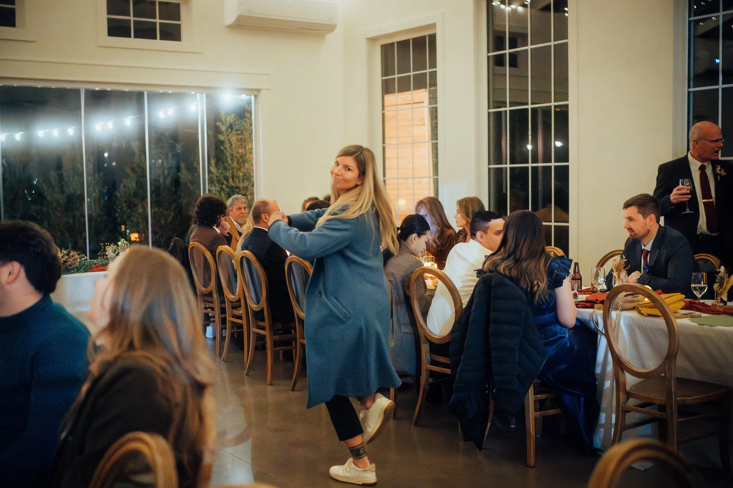 People attending a formal dinner or celebration in a well-lit room with large windows, seated at round tables with tableware, candles, and decorative centerpieces, with a woman in a blue coat standing and smiling at the camera.