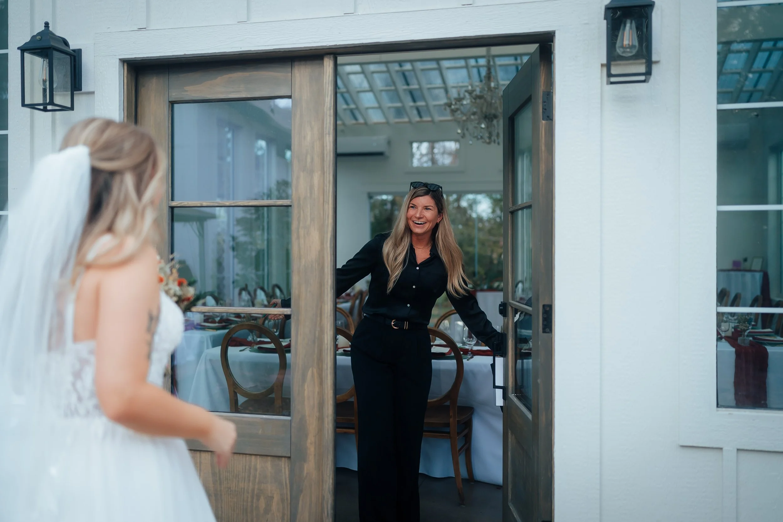 A woman in a black outfit smiling and holding the door open for a bride in a white wedding dress at a banquet hall.