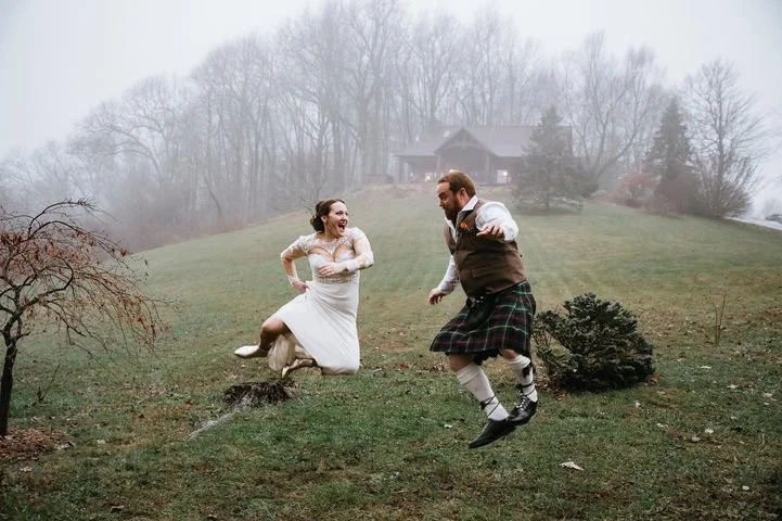 A man and woman in traditional Scottish attire happily jumping and dancing outdoors on a foggy day with a house and trees in the background.