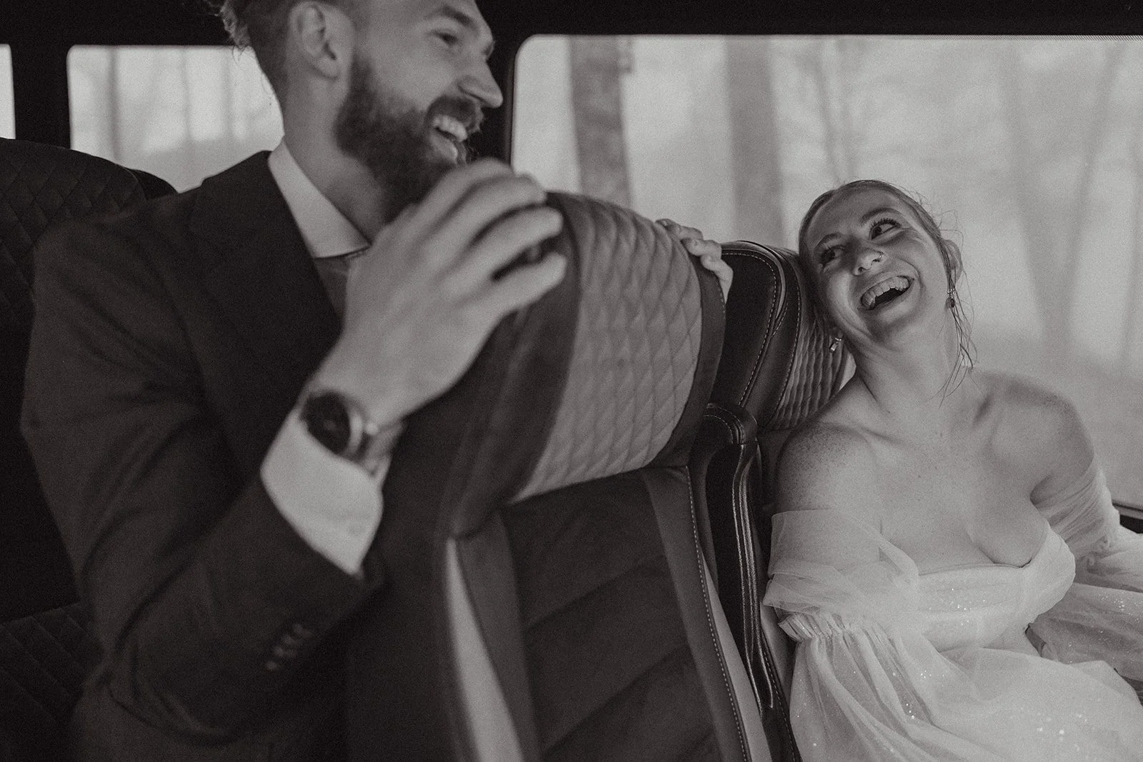 A man and woman are sitting on a bus, smiling and laughing together. The woman is dressed in a wedding gown, and the man is wearing a suit.