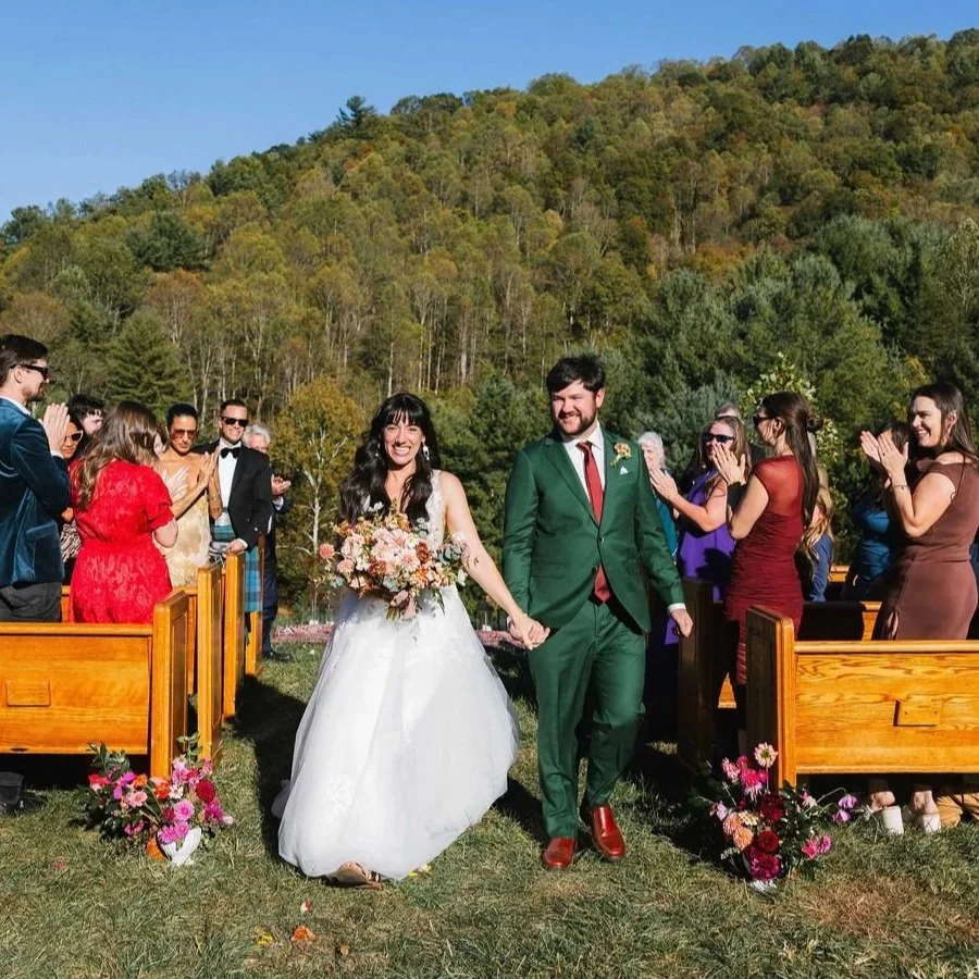 Bride and groom walking hand in hand after their wedding ceremony outdoors, with guests clapping and smiling on either side, green trees and hills in the background.