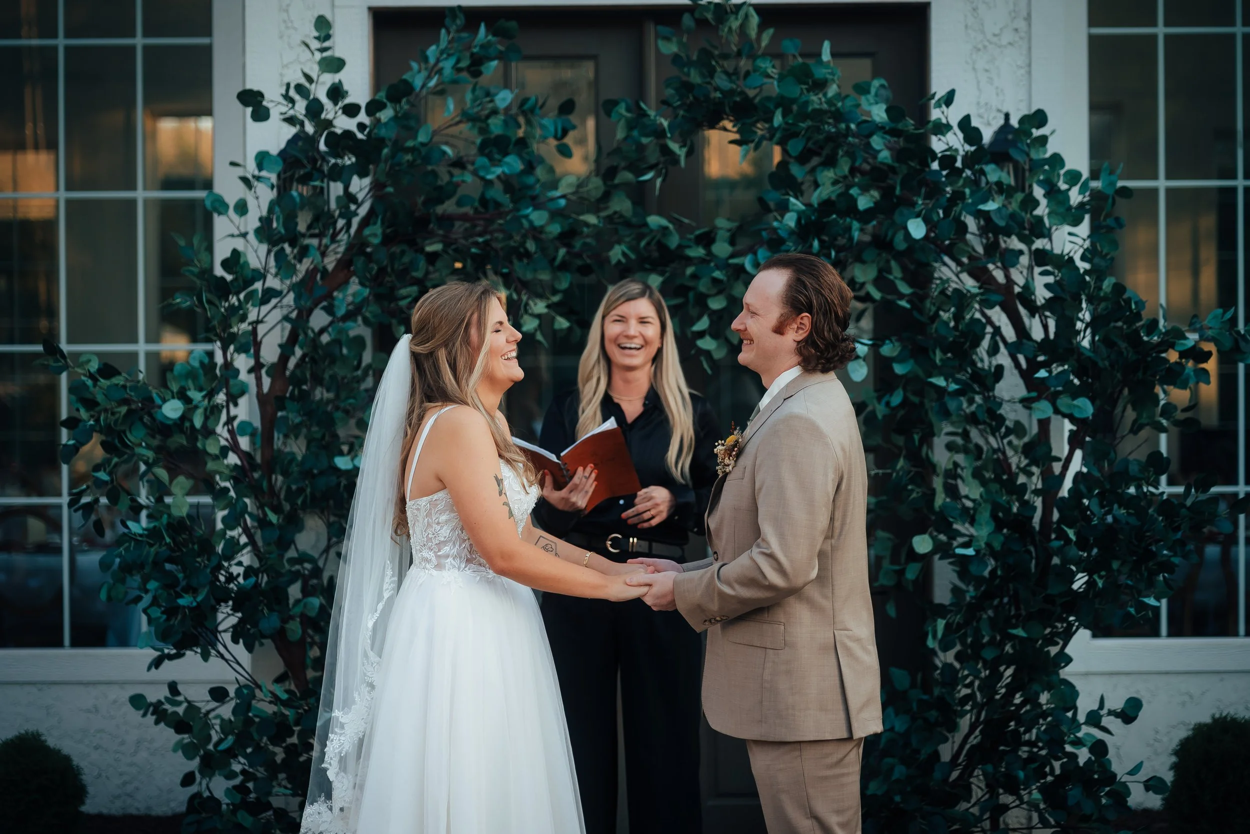 A couple getting married, holding hands and smiling at each other, with a female officiant in a black outfit standing behind them, outdoors in front of a building with a leafy arch.