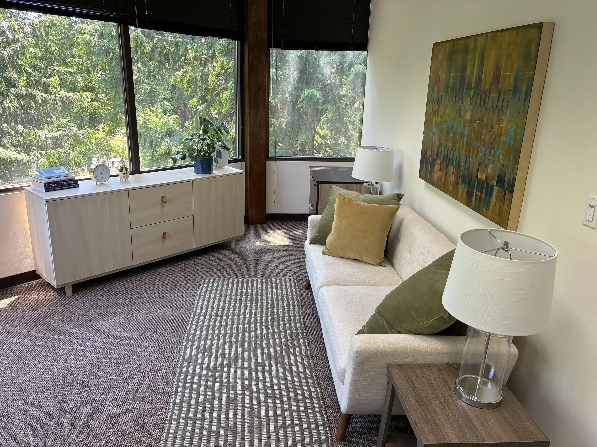 A cozy corner in a room with large windows overlooking trees, a white sideboard with books, a clock, and small decorative items, a potted plant, a beige sofa with green and tan pillows, a wooden side table with a glass lamp, and a large colorful painting on the wall.