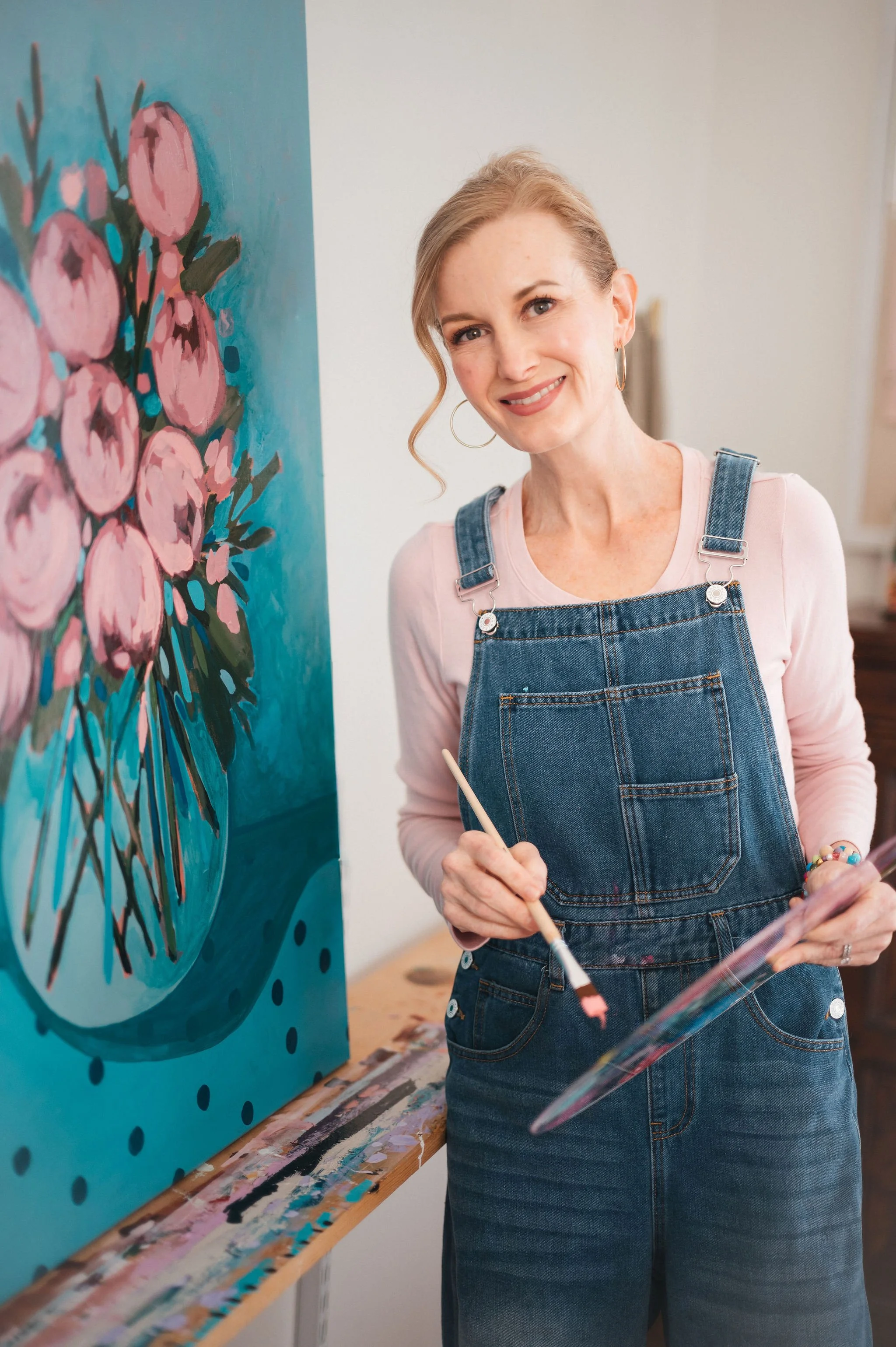 a woman in a pink shirt and jean overalls is standing next to a large floral painting, holding a paintbrush dipped in pink paint