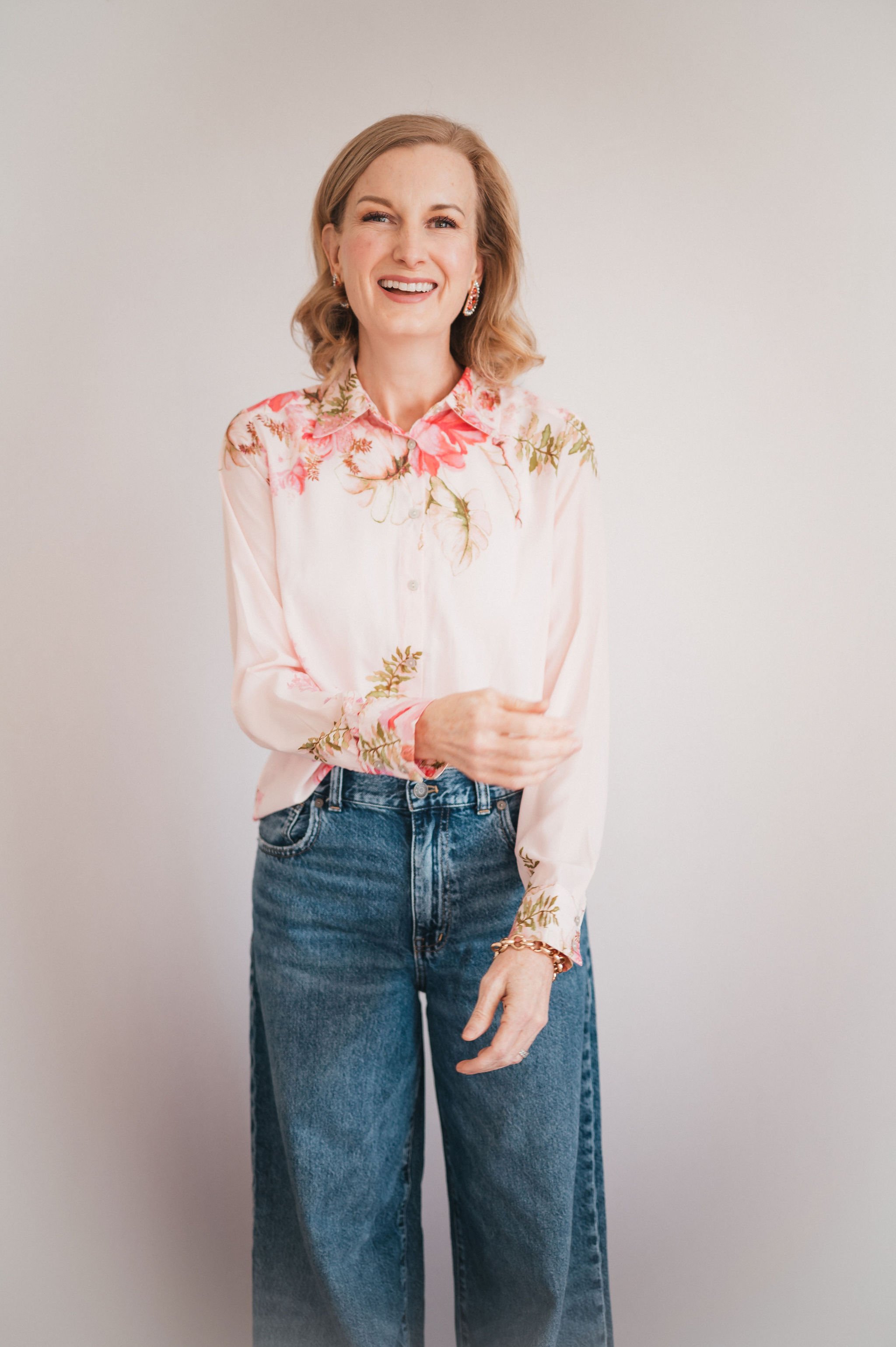 a woman in jeans and a floral pink button up is smiling widely and standing in front of a white wall