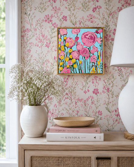 a painting of yellow and pink flowers hangs on a pink wallpapered wall, above a dresser with books and a lamp on top