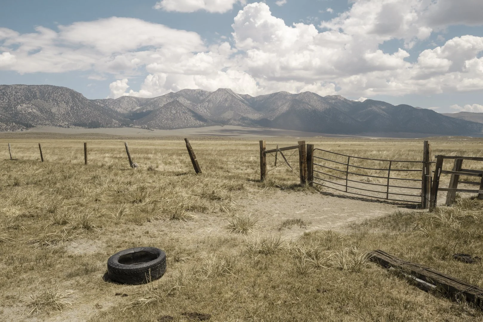 Open rural field with a broken wooden fence and a discarded tire in the foreground, mountains in the distance, partly cloudy sky.