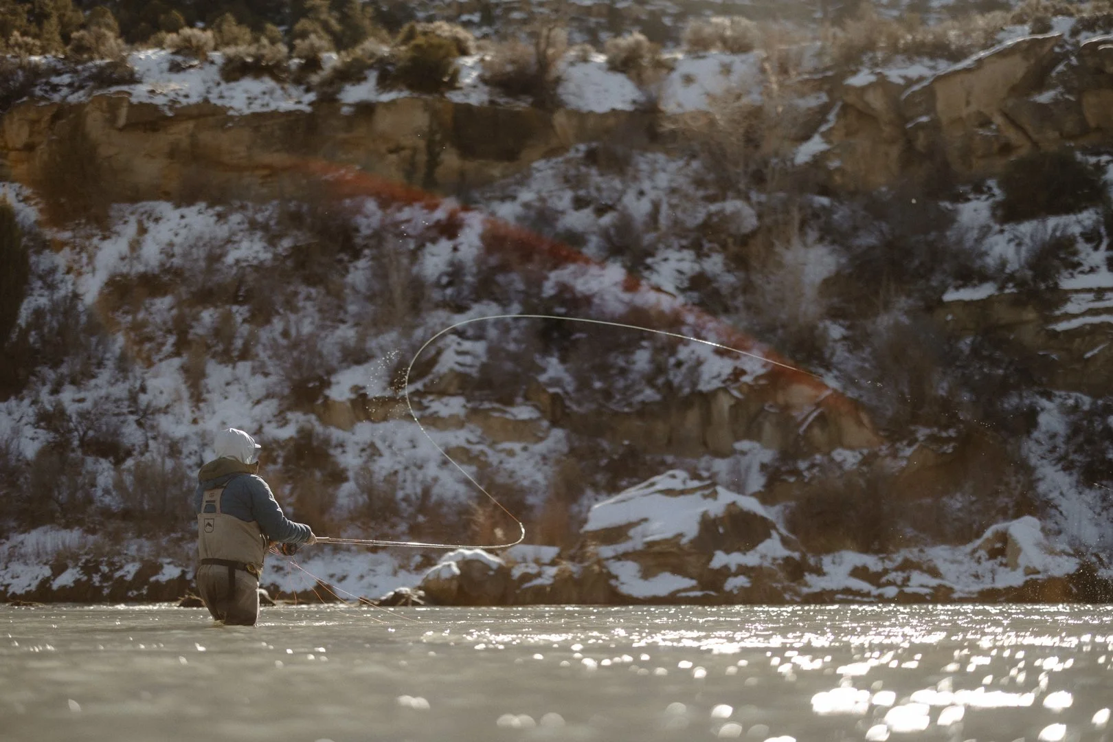 A person standing in a frozen river fishing with a rod, with snow-covered rocks and hillside in the background.