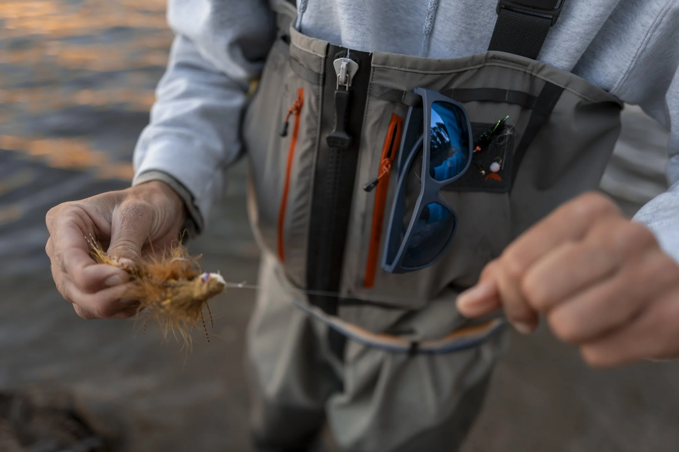 Person tying fishing line with a fishing lure, wearing a fishing vest with sunglasses attached, near a body of water at sunset.