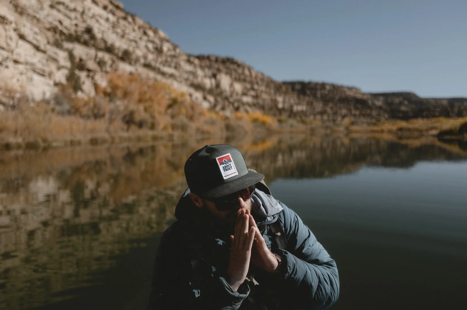 A man wearing a black cap, sunglasses, and a blue jacket, standing by a calm lake with mountains in the background, during autumn.
