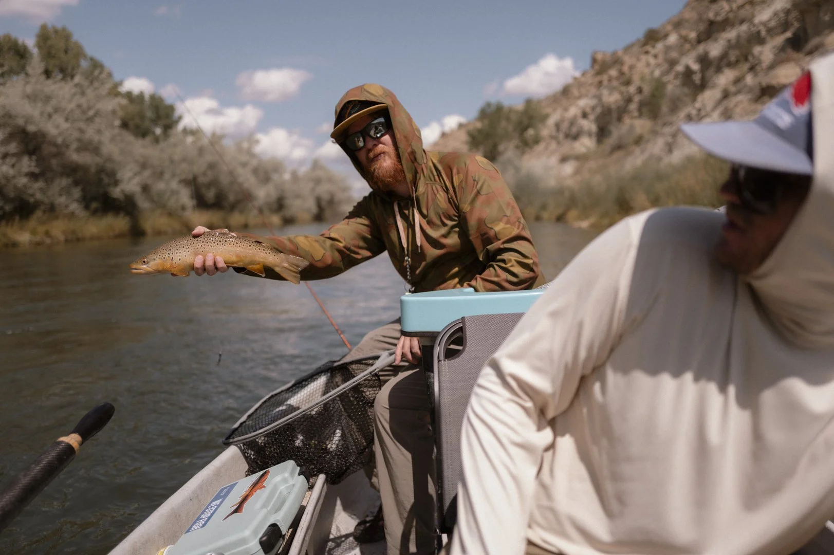 Two men fishing from a boat on a river, one holding a brown trout. The man holding the fish is wearing camouflage jacket, sunglasses, and a hat with a hood, and the other man, wearing a white hoodie and cap, is partially visible.