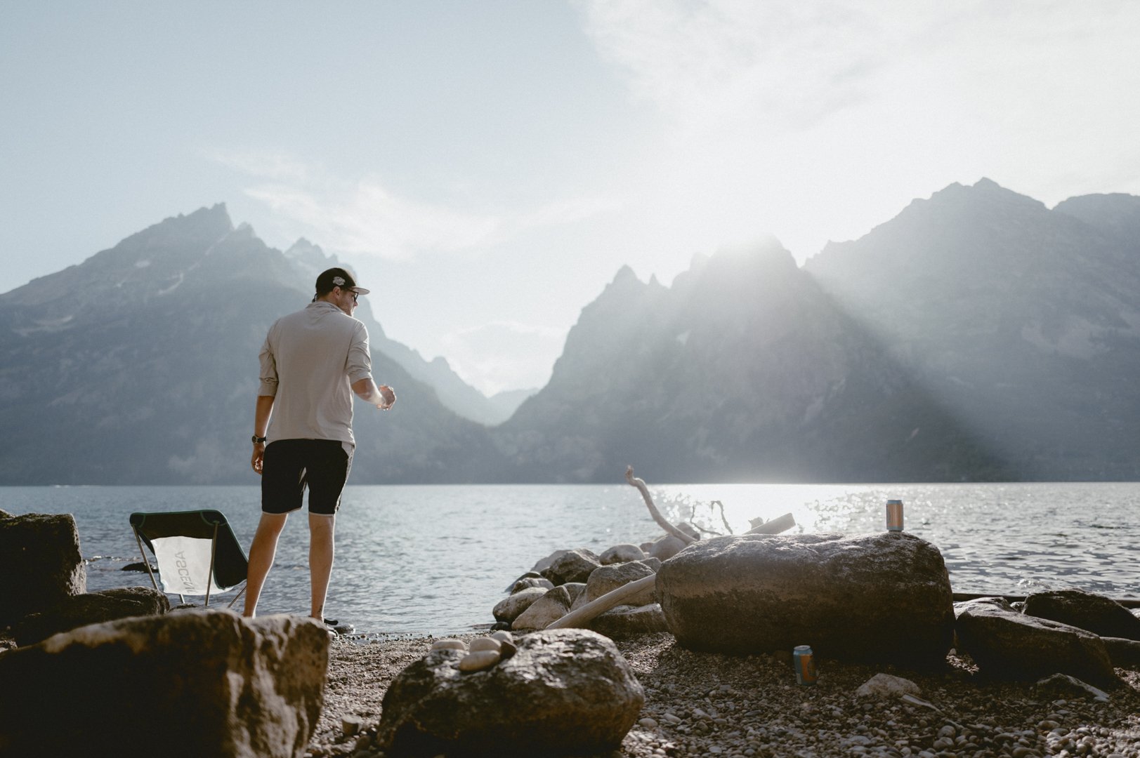 A man standing by a lake with mountains in the background, holding a drink and looking at the scenery.