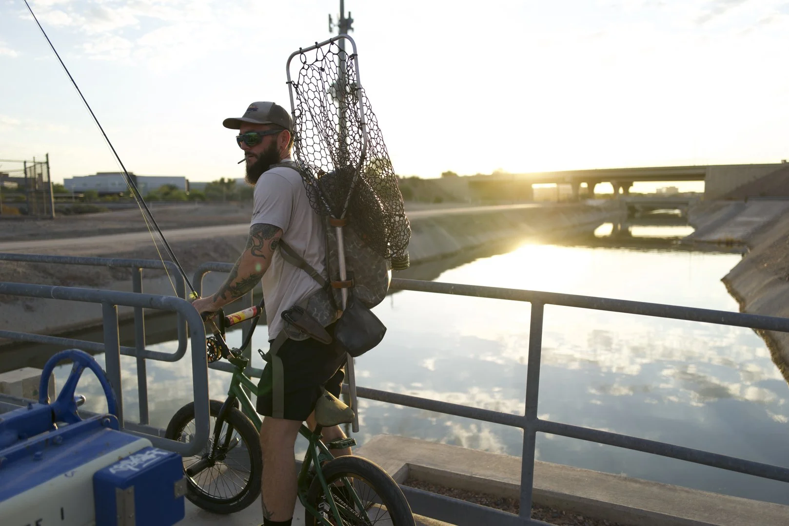 A man with tattoos, wearing sunglasses, a cap, and a gray t-shirt, standing with a bicycle by a waterway at sunset, carrying a backpack and a fishing net.