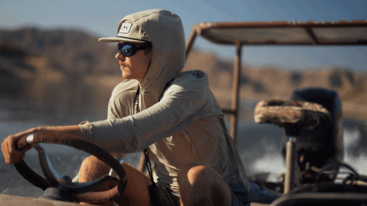 A man wearing sunglasses, a hoodie, and a cap driving a boat in a body of water with mountainous terrain in the background.