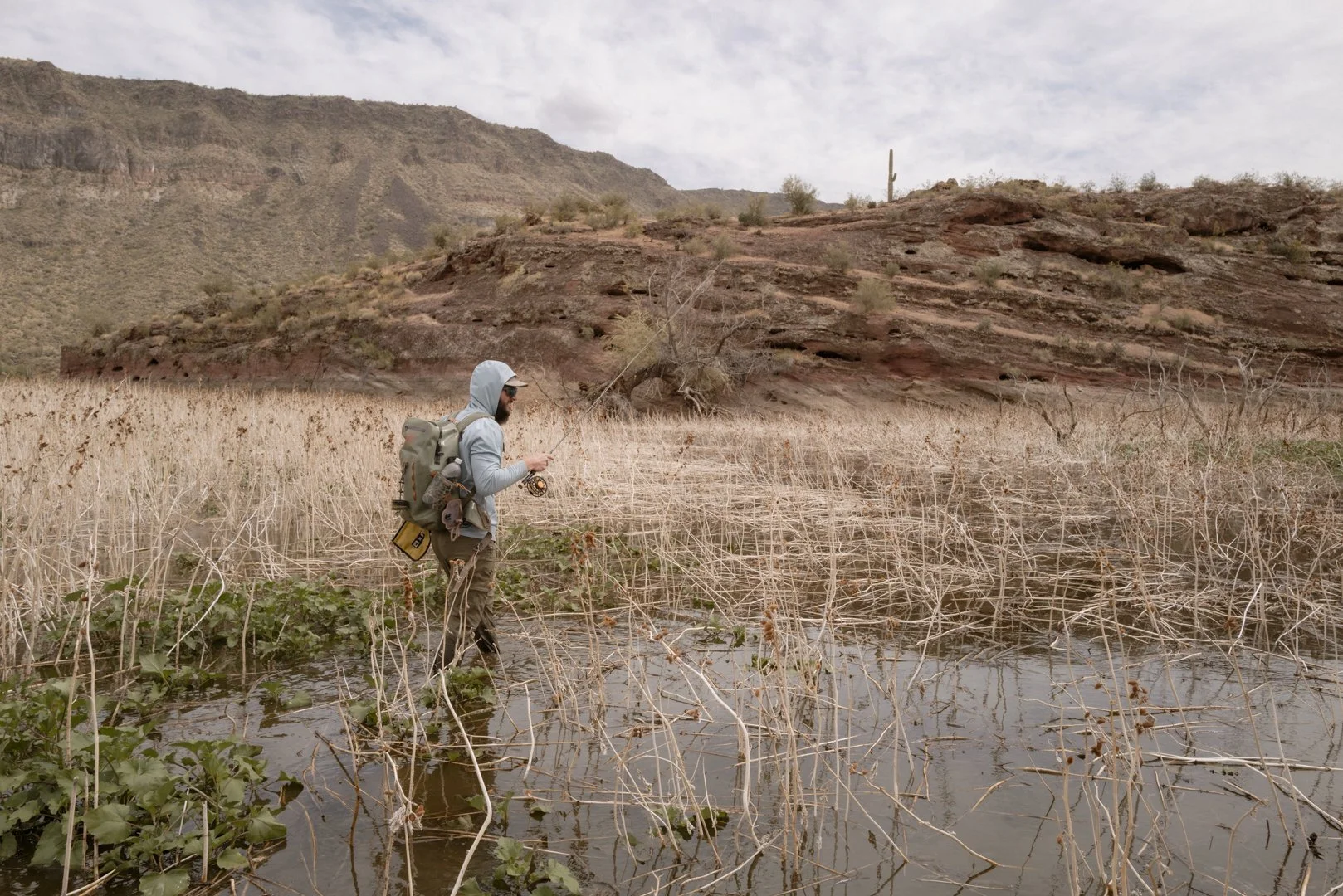 A man in outdoor clothing, including a hooded jacket and sunglasses, walking through a wet marshy area with a backpack, holding a fishing rod in a desert landscape with brown mountains and sparse vegetation.