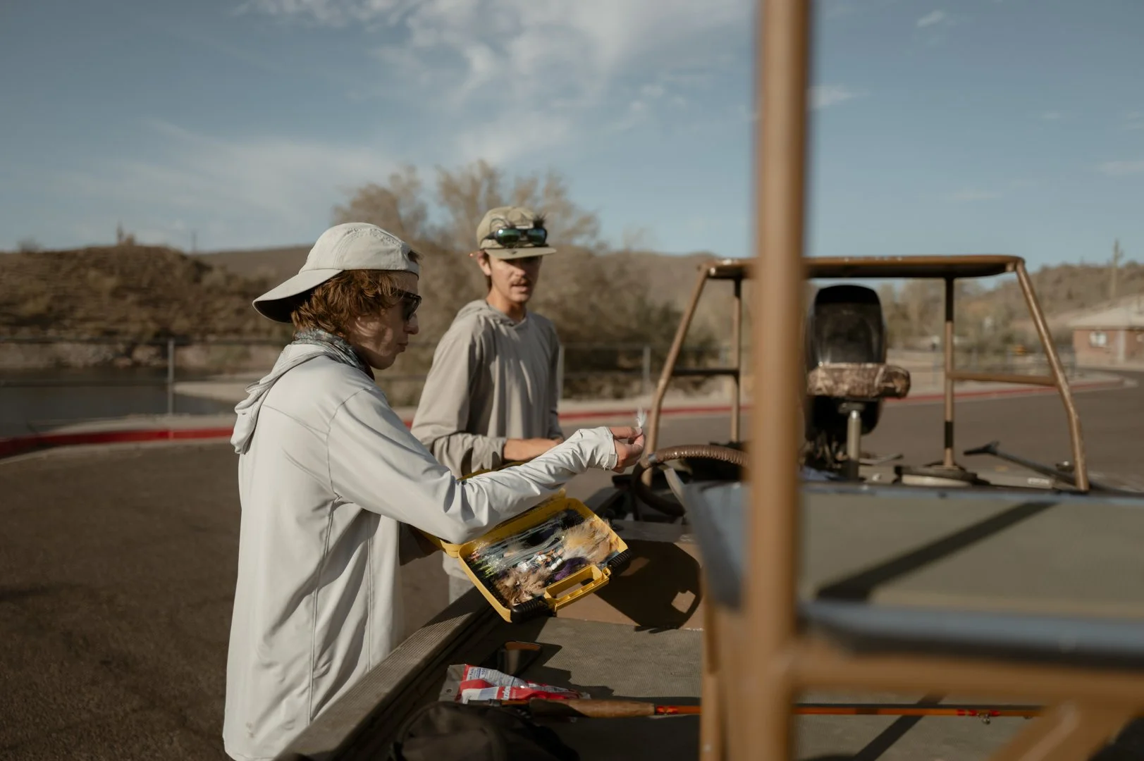 Two young men stand beside a golf cart on a golf course during daytime. One of them, wearing a beige cap backwards, sunglasses, and a white jacket, appears to be preparing or handling some small object, while the other, wearing a beige hat with binoc