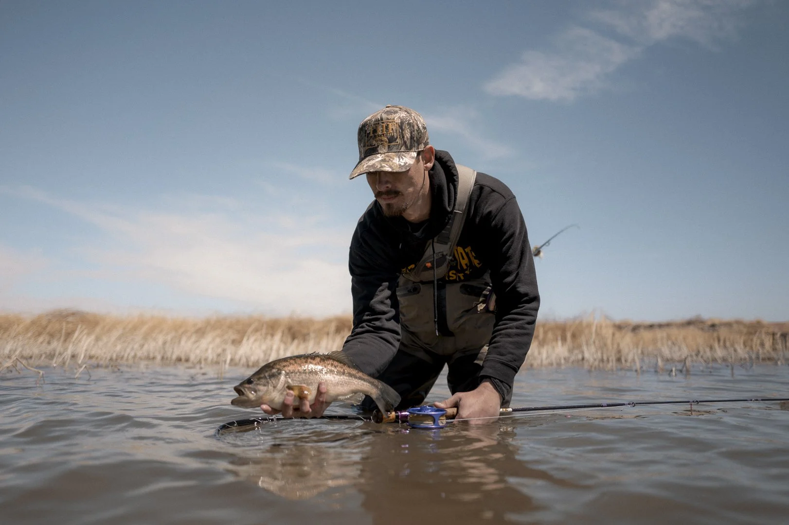 Man holding a fish in a shallow body of water, with a fishing rod on the water surface and dry grass on the shoreline, under a partly cloudy sky.