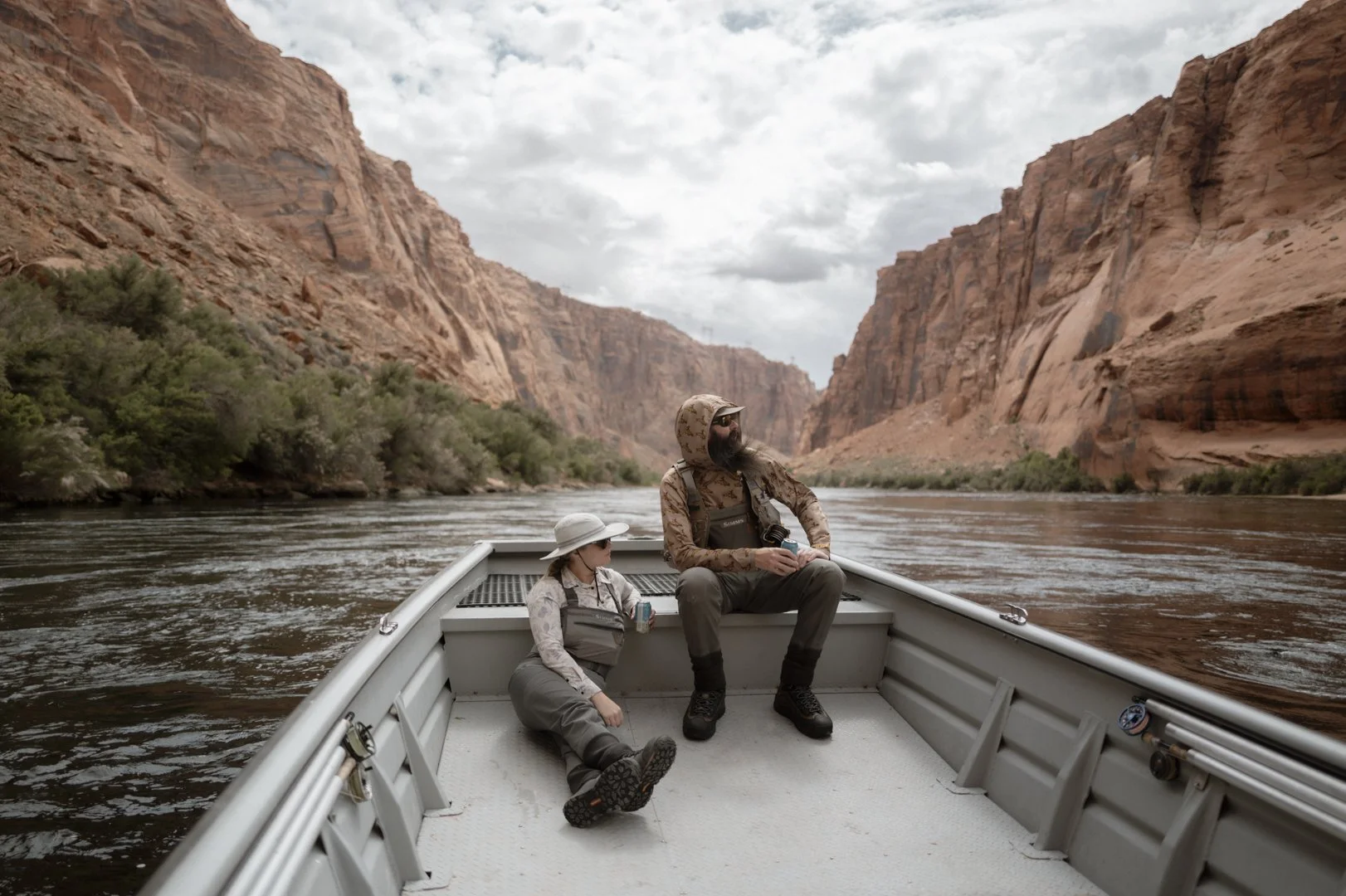 A man and a child sitting on a boat in a canyon with a river, red rock cliffs, and cloudy sky in the background.