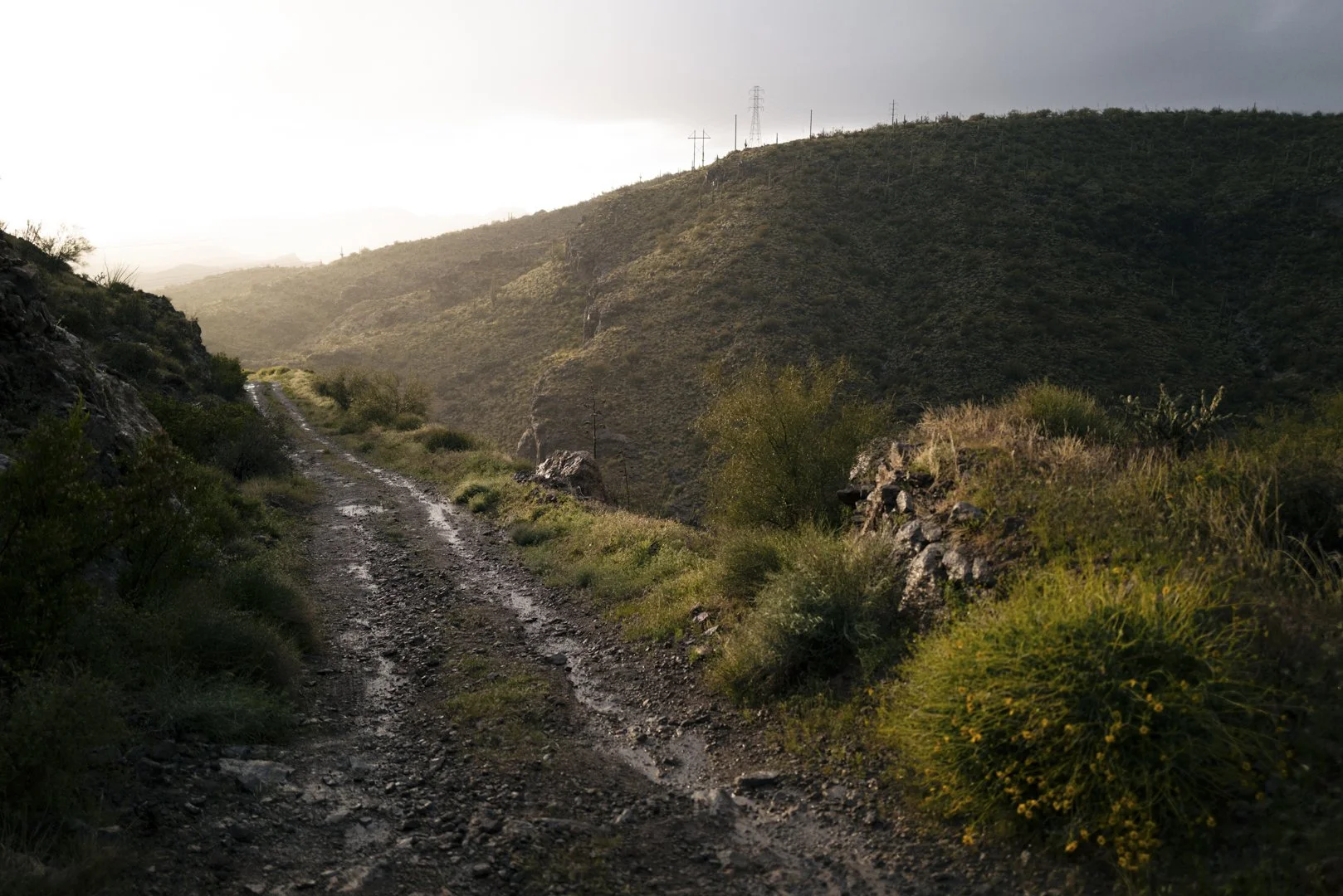 A rugged dirt trail winding through a remote hilly landscape with sparse vegetation and shrubs, under a cloudy sky with sunlight breaking through.