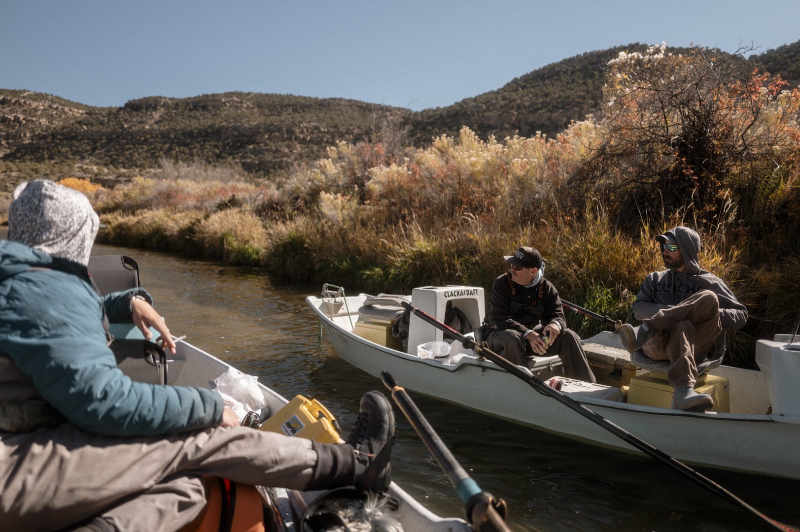 Four people on two small boats fishing on a river during autumn, with colorful foliage and hills in the background.