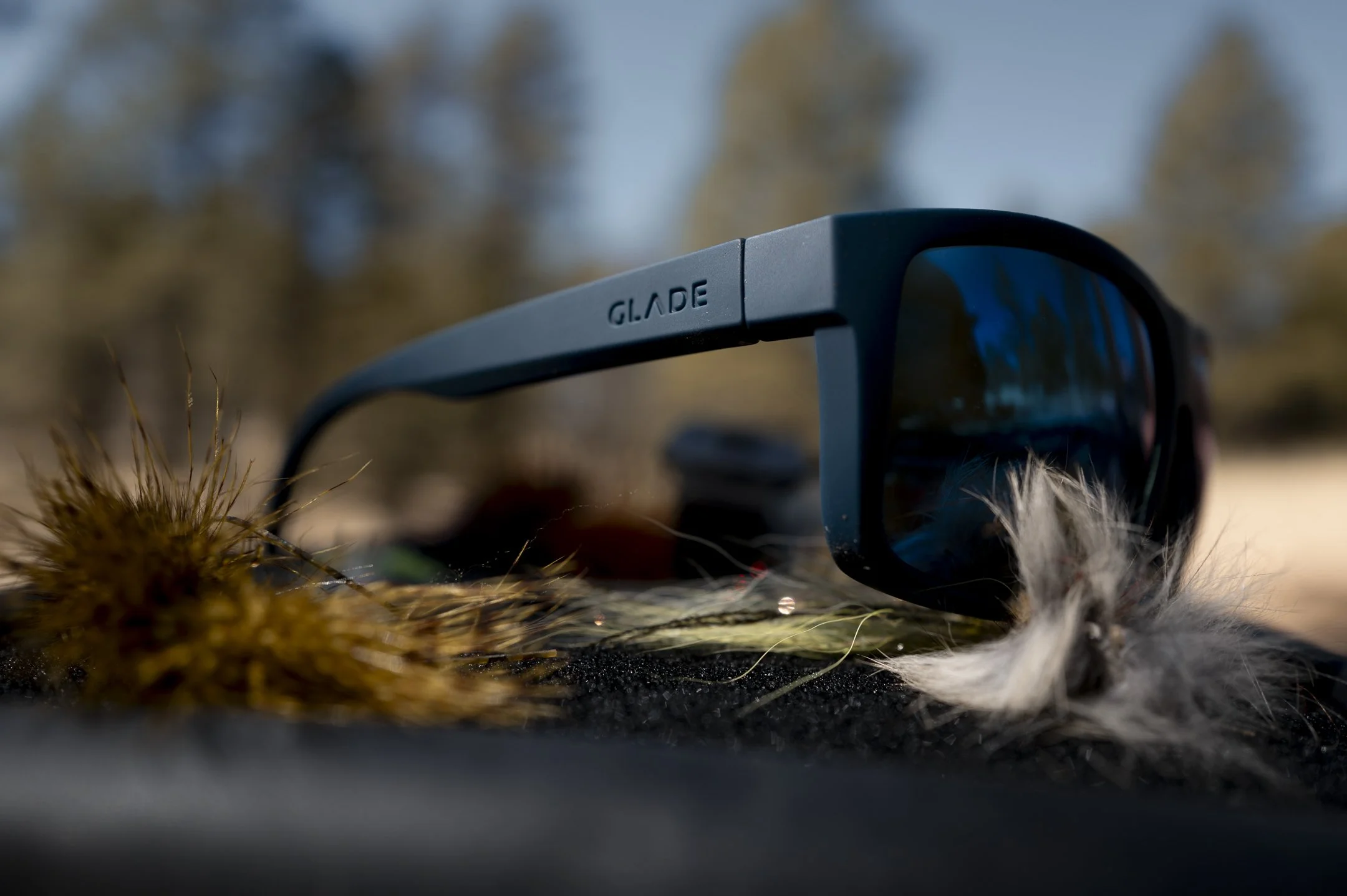 Black GLARE sunglasses resting on a surface with some dried grass and fur, outdoors with trees in the background.