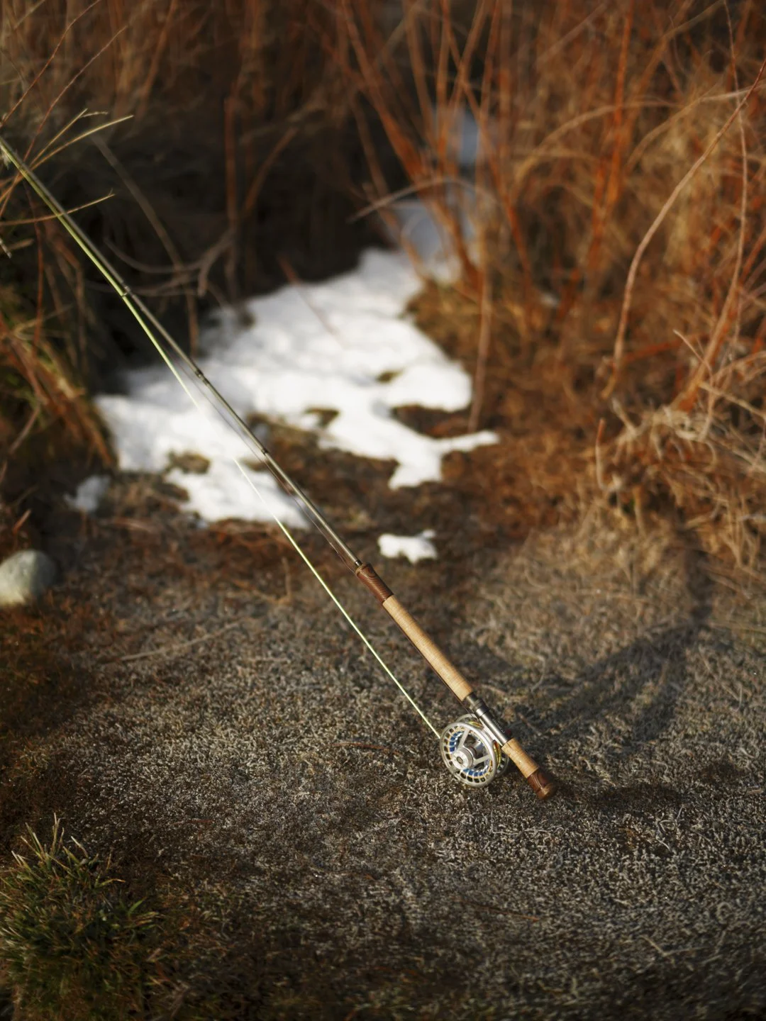 A fishing rod lying on a rocky ground near a small stream with snow patches, surrounded by dry grass and shrubs.