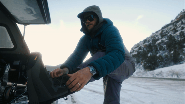 A man dressed in winter clothing, including a blue jacket, hood, and sunglasses, bending down to adjust equipment on a snow-covered road with snowy hills in the background.