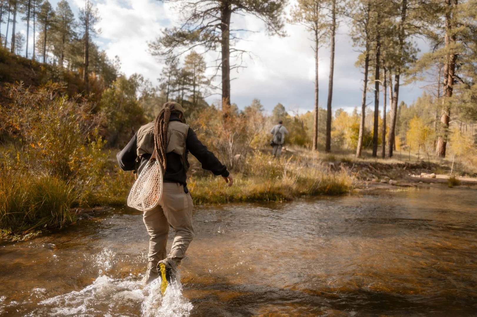 Two people fishing in a creek through a forest during autumn.