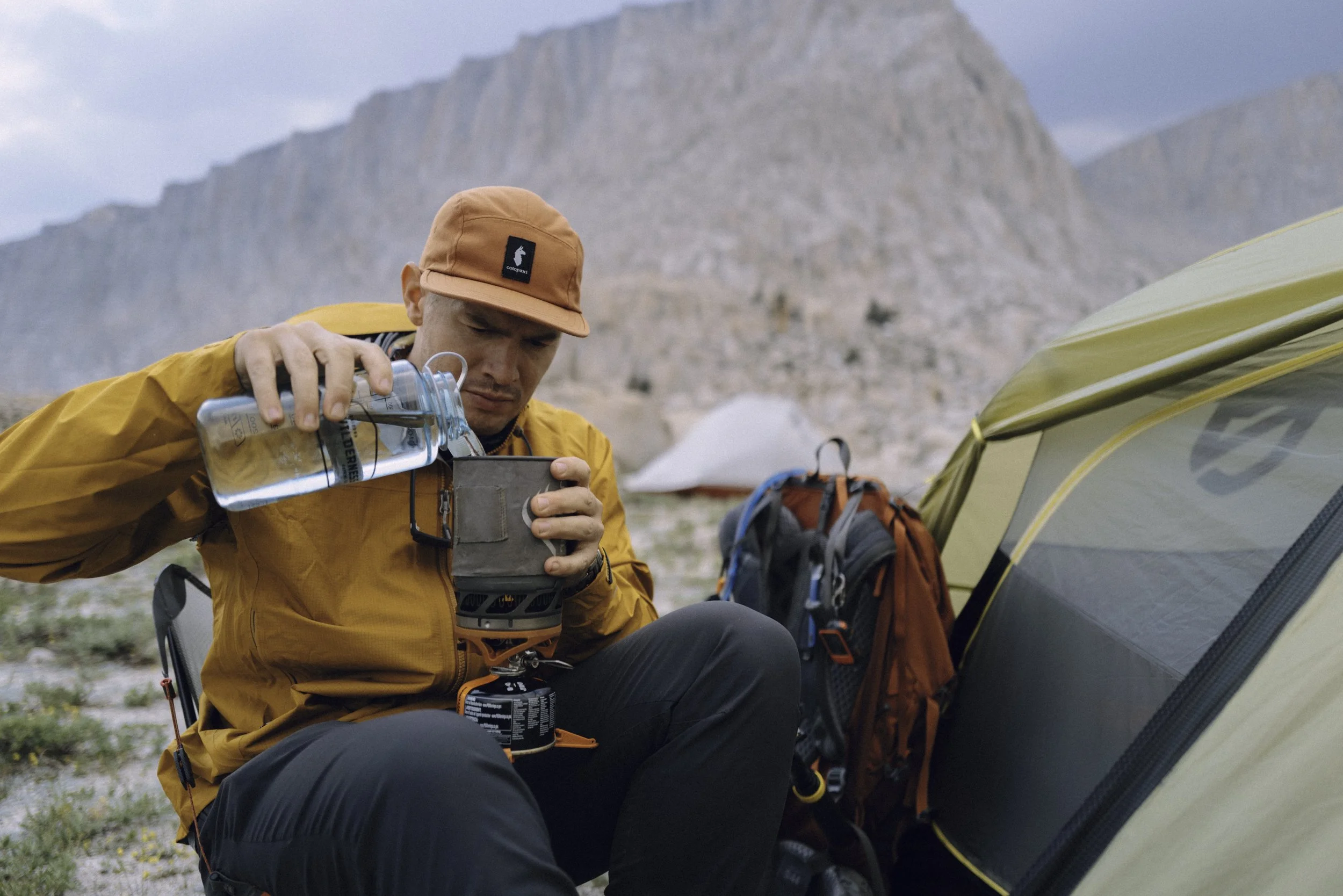 A man in an orange cap and yellow jacket prepares water using a portable stove next to a tent and backpack in a mountainous outdoor setting.