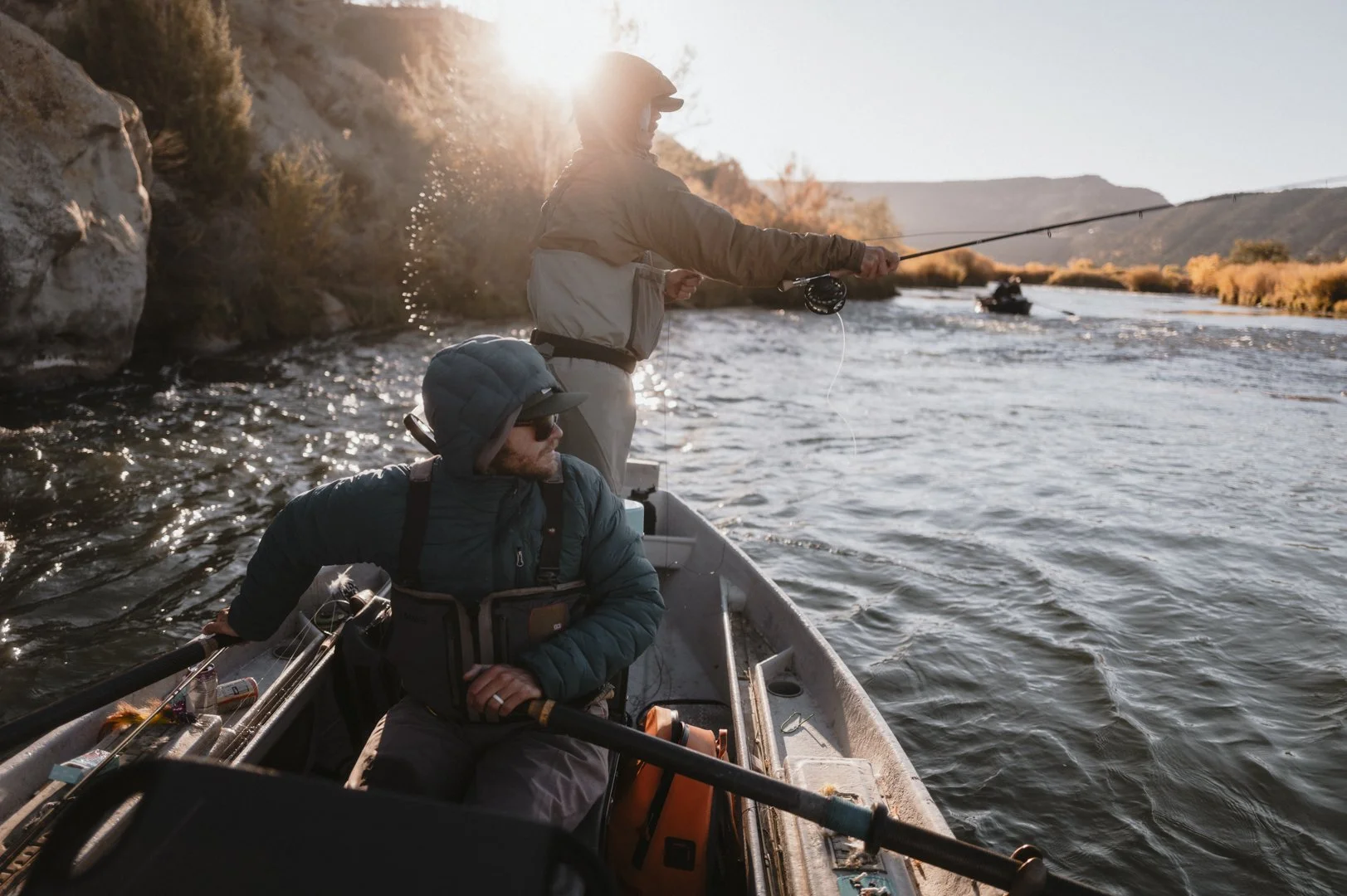 Two men fishing from a boat on a river at sunset, with rolling hills and trees in the background.