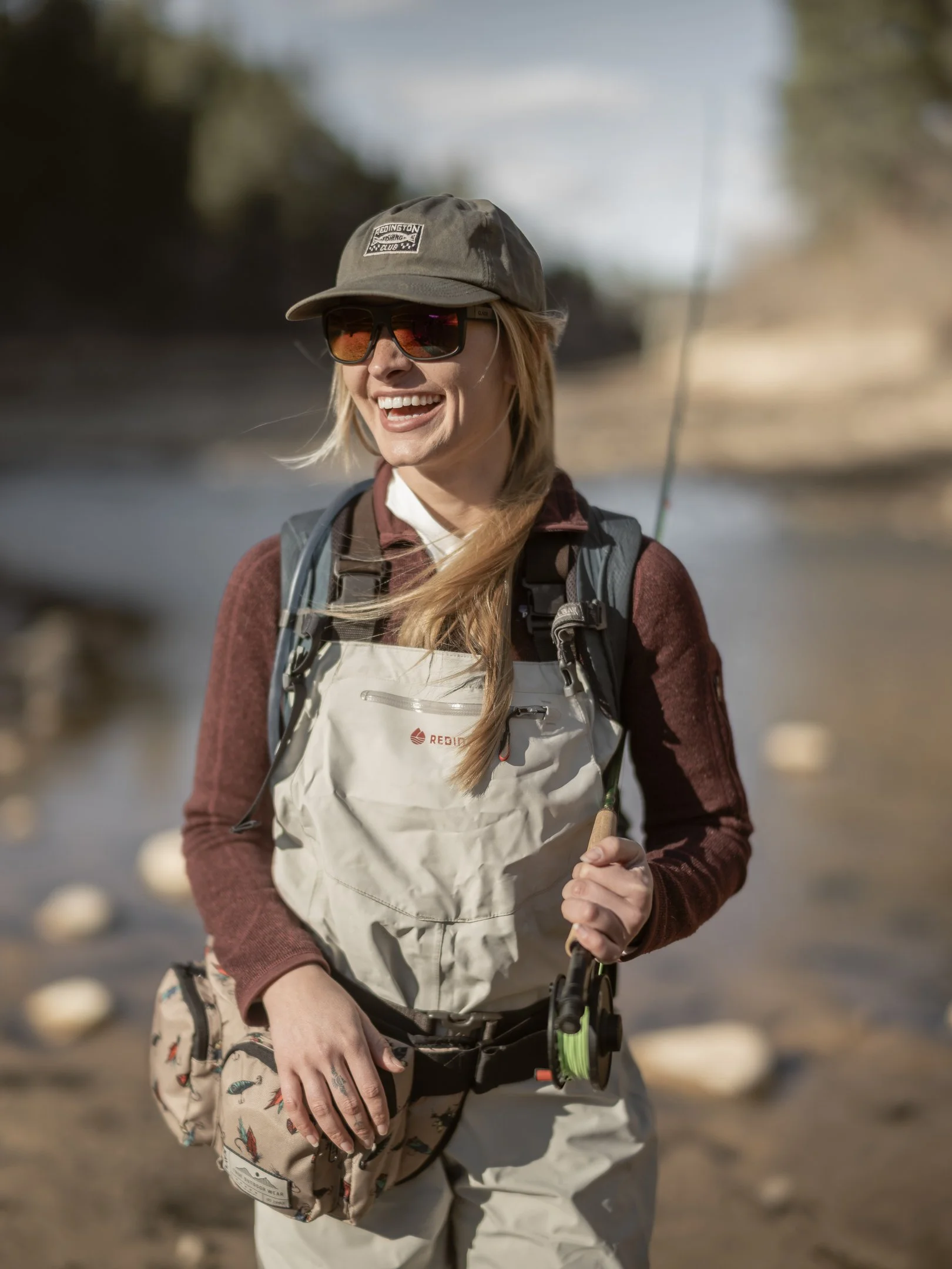 A smiling woman outdoors wearing sunglasses, a hat, a backpack, and fishing gear, holding a fishing rod.
