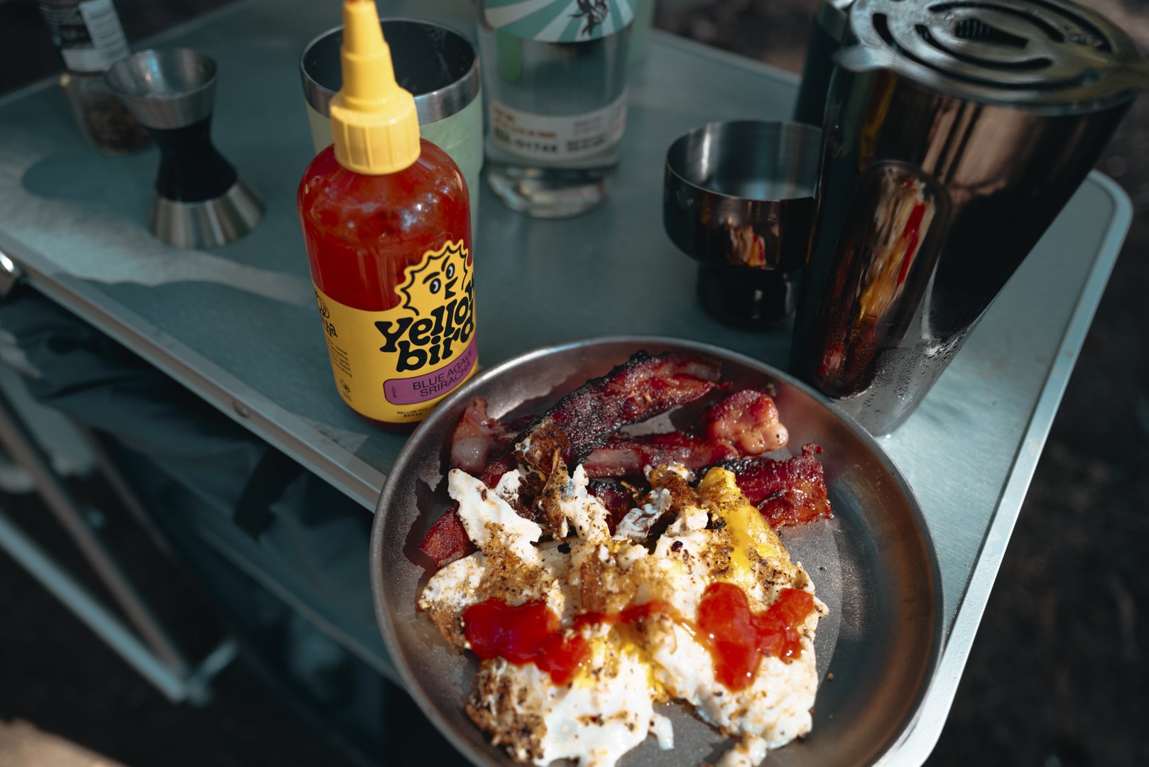 A camping table with a stainless steel plate of cooked breakfast, including bacon, eggs with ketchup, and sausage links. There is a bottle of Yellowbird hot sauce, a glass of soda, and various cups and containers visible on the table.
