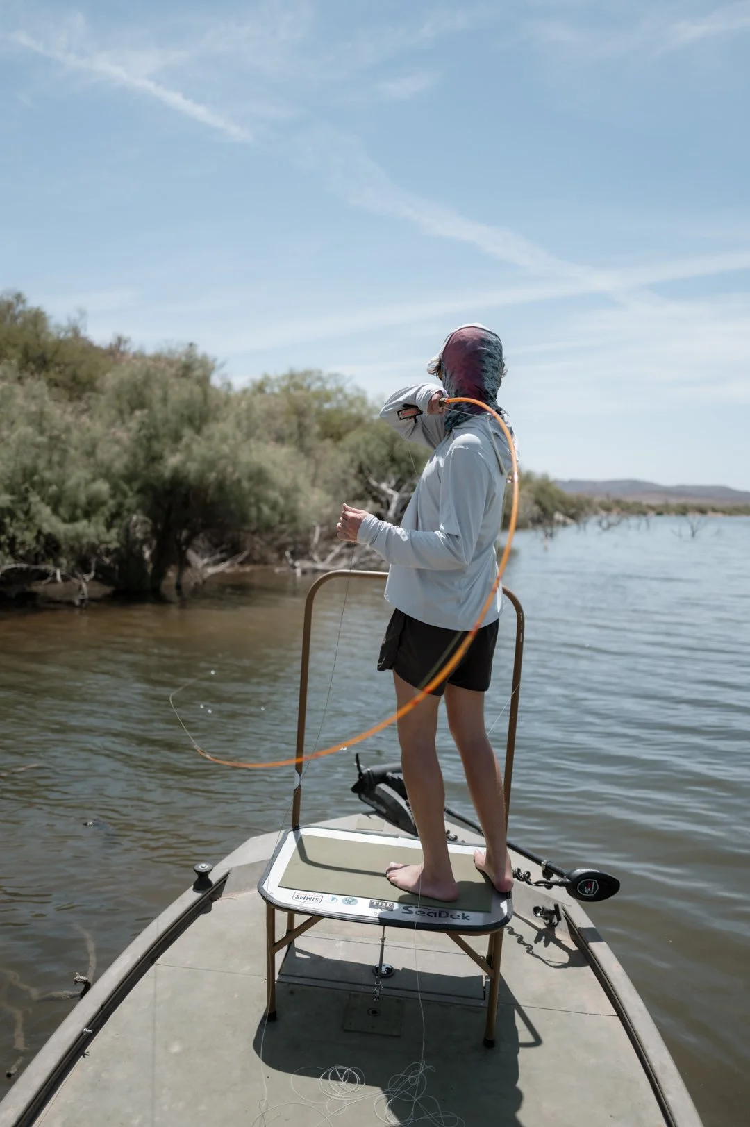 A person wearing a hoodie, shorts, and no shoes stands on a small table on a boat, fly-fishing in a river with trees and blue sky in the background.