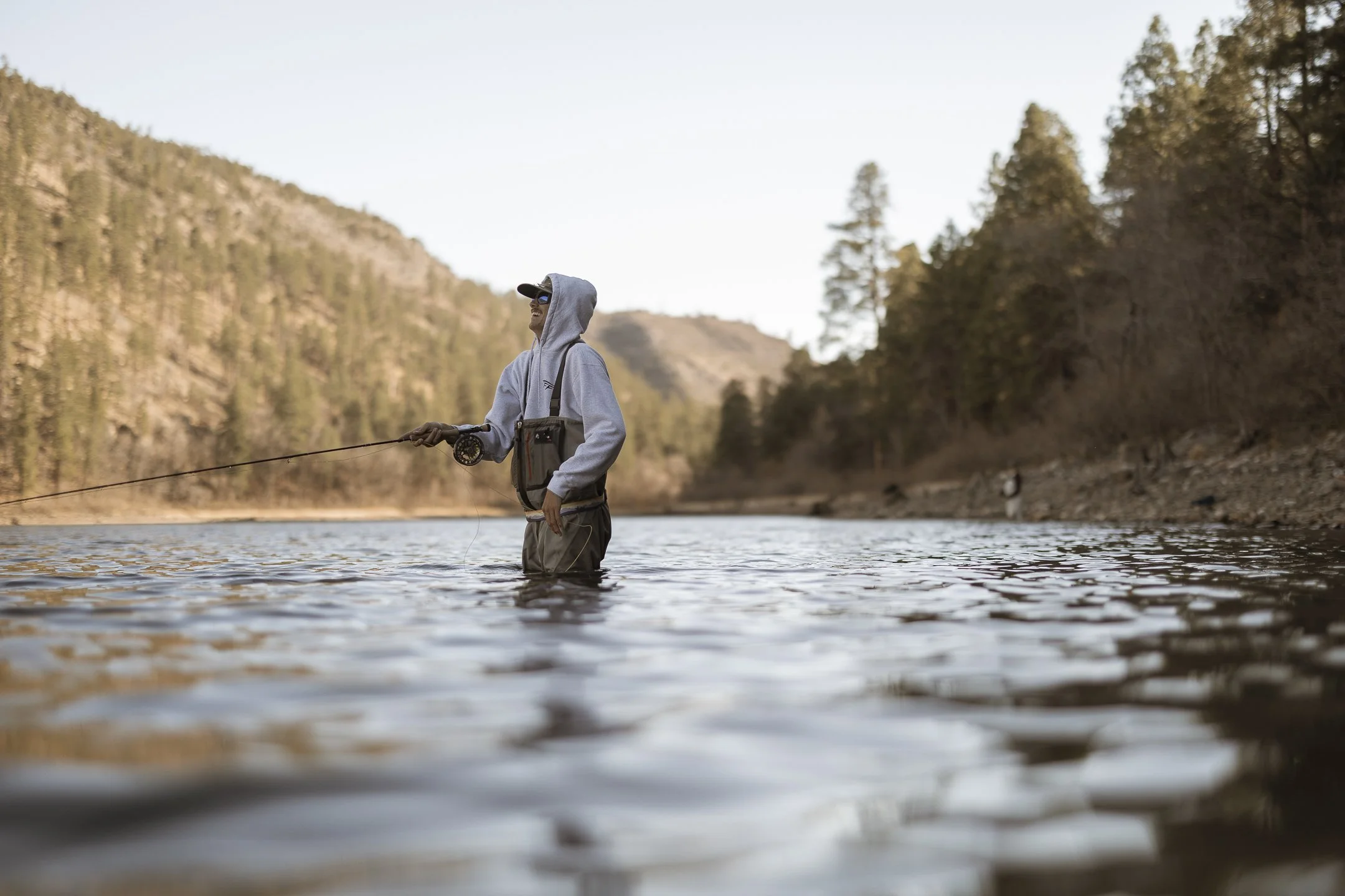 Person fishing in a river surrounded by trees and mountains.