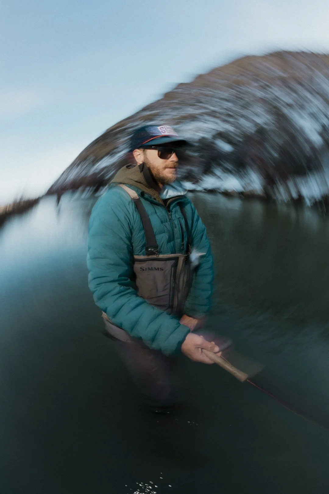 Man fishing in a river, wearing a blue jacket, sunglasses, a cap, and a life vest, with water splashing around him.