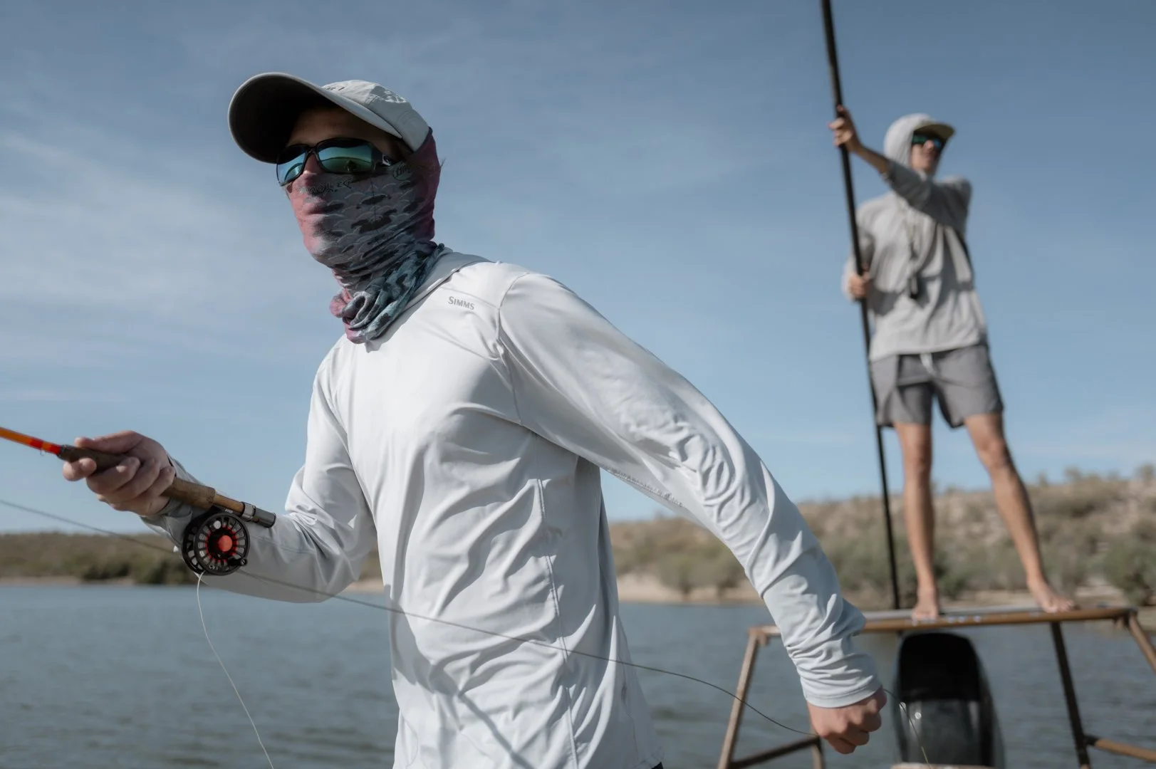 Two men fishing on a boat in a calm body of water under a clear blue sky. One man in the foreground is wearing sunglasses, a face covering, a light-colored hat and long-sleeved shirt, holding a fishing rod. The other man in the background is standing