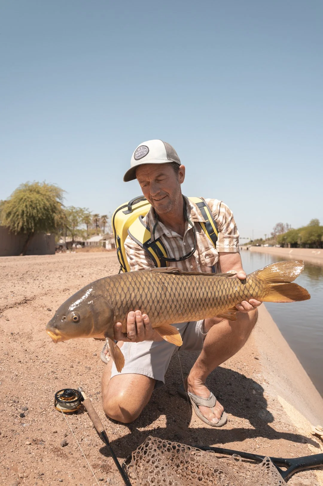 A man kneeling on the ground holding a large fish with a stream and some trees in the background.