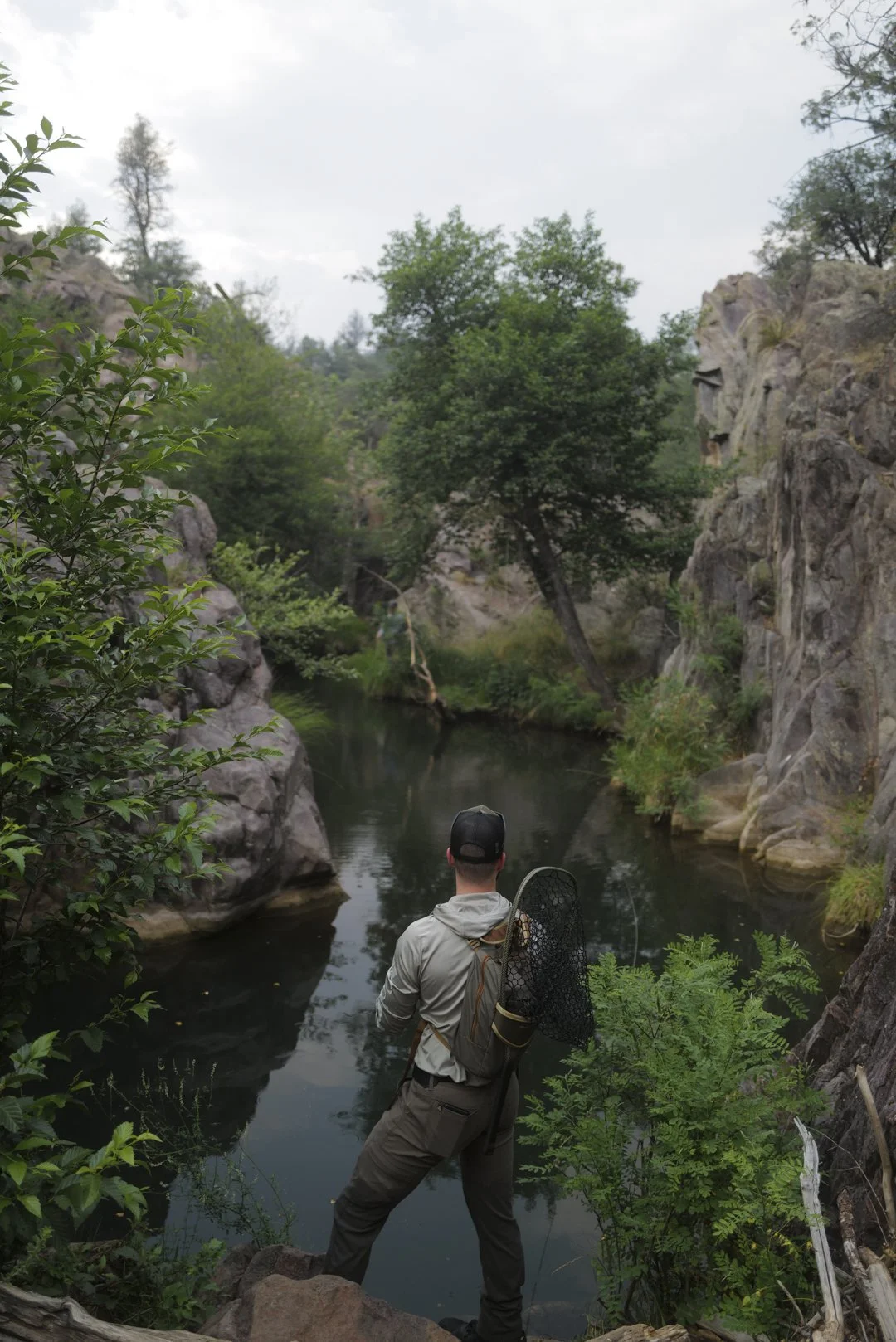 A man fishing in a tranquil river surrounded by rocks and trees in a natural setting.