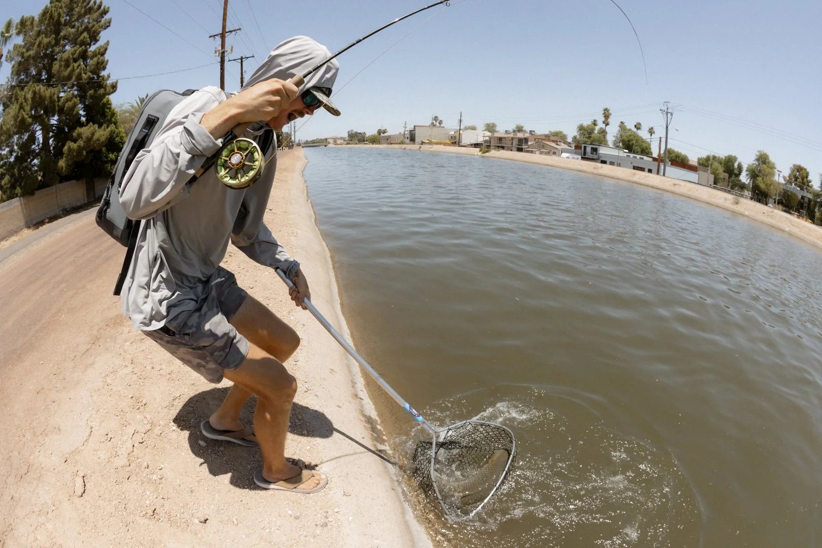 Man fishing in a pond, holding a fishing rod, wearing sunglasses, hoodie, shorts, and flip-flops, standing on a sandy shore with houses and power lines in the background.