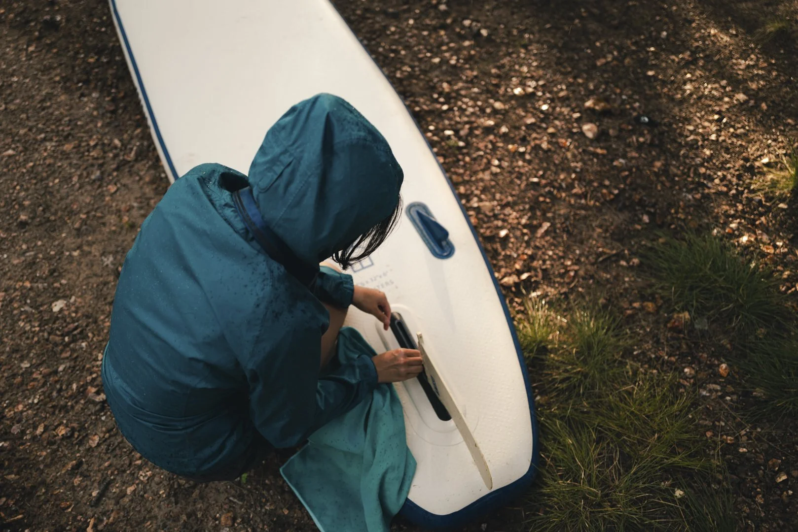Person in teal rain jacket with hood sitting on ground next to a white paddleboard, writing in a notebook, surrounded by damp rocks and grass.