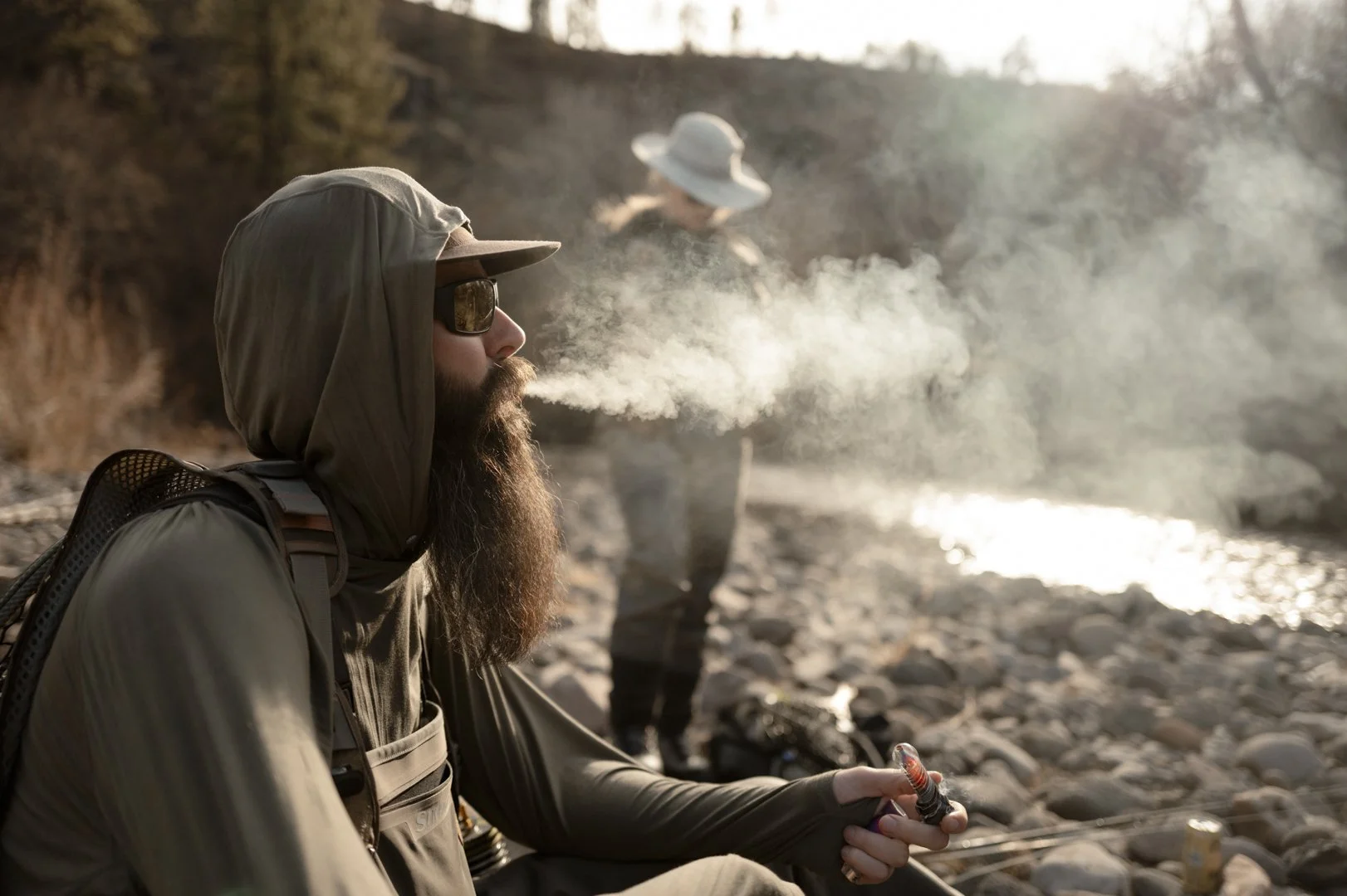 A man with a beard wearing a hoodie, sunglasses, and a cap is sitting on rocks near a river, blowing smoke from a pipe. Another person in the background is also near the river, wearing a hat.