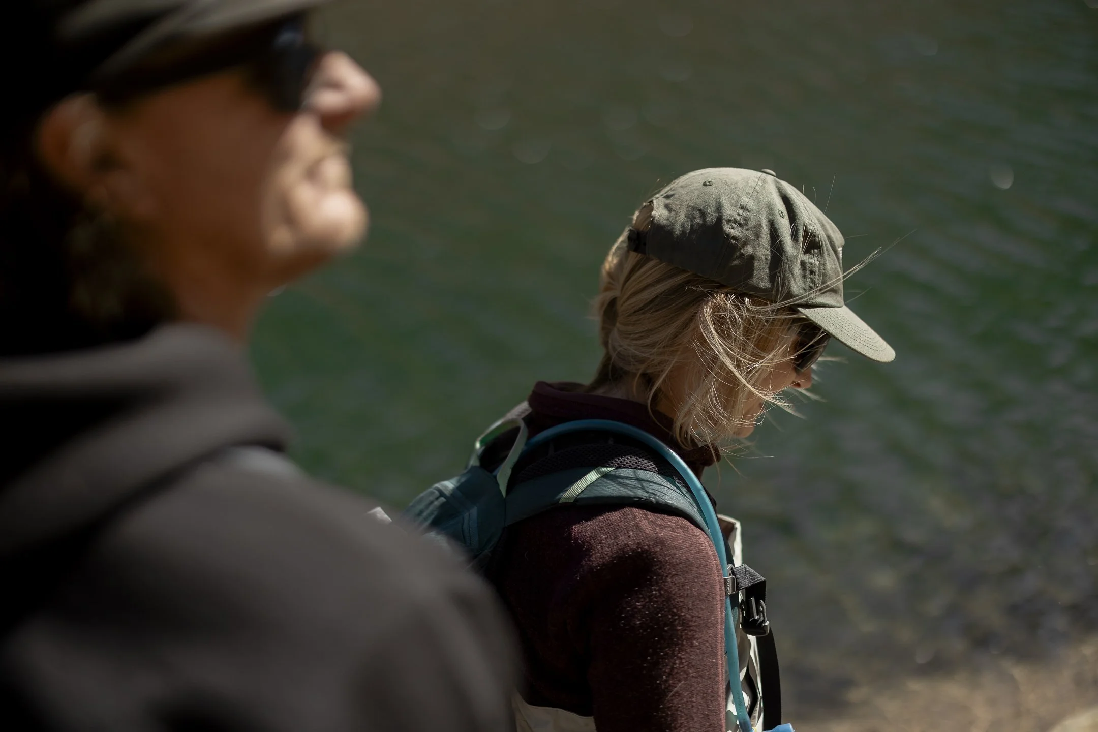 Two women near a body of water, one is in the foreground and blurred, the other is in the background wearing a hat.