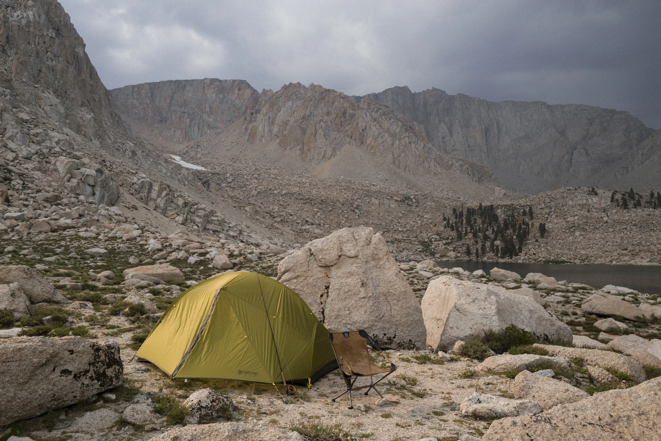 A green Nemo Equipment camping tent and a folding chair set up on rocky terrain near a lake, with mountains and cloudy sky in the background.